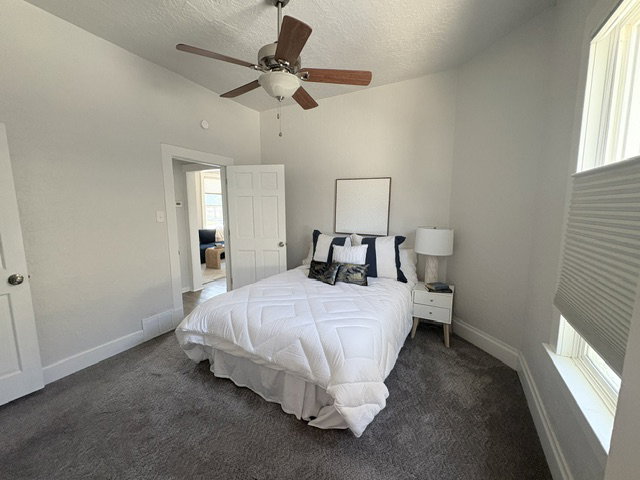 Carpeted room featuring a ceiling fan with wood-finish blades, a white paneled door, and a window with cellular shades