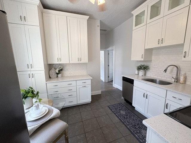 Modern kitchen featuring white shaker cabinetry, stainless steel dishwasher, white subway tile backsplash, light-toned countertops, and dark tile flooring