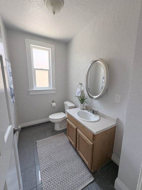 Bathroom featuring a light wood-finish vanity with a white countertop and integrated sink, a framed oval mirror, a white toilet, and gray tiled flooring