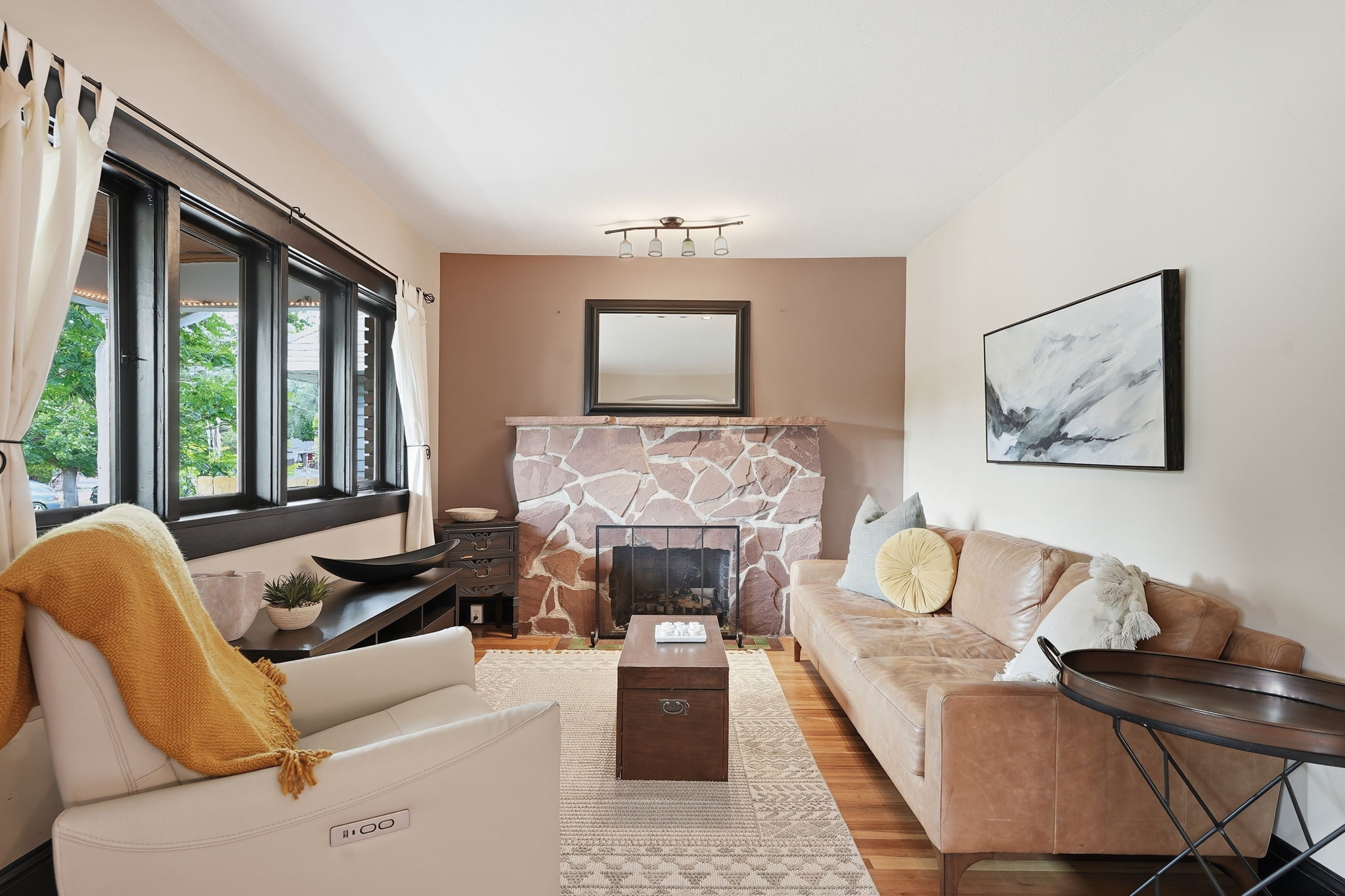 Living area featuring a stone fireplace with a metal screen, wood-finish flooring, and multiple windows with dark trim and white curtains