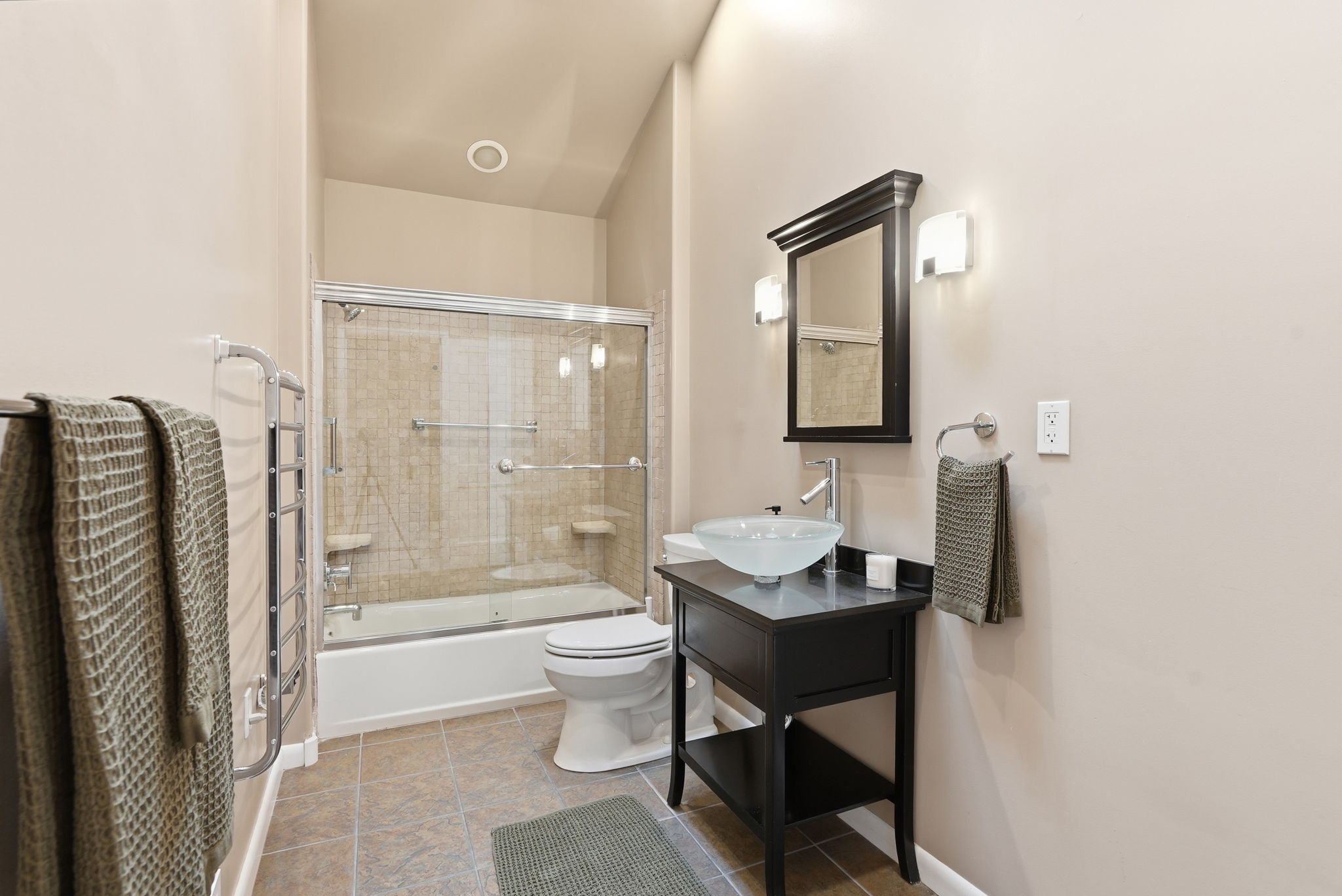 Bathroom featuring a tub/shower combination with sliding glass doors and tiled surround, a black vanity with a vessel sink, a mirrored medicine cabinet, and sconce lighting