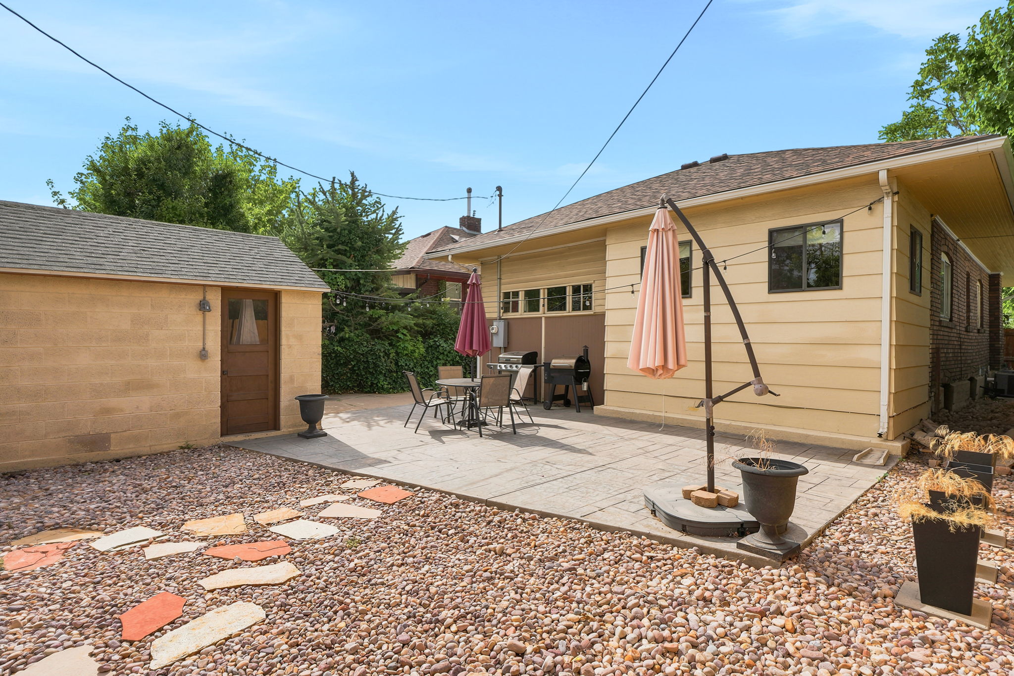 Expansive gravel yard featuring a rectangular stone patio, a detached storage building with a wood door, and a light-colored exterior wall on the main residence