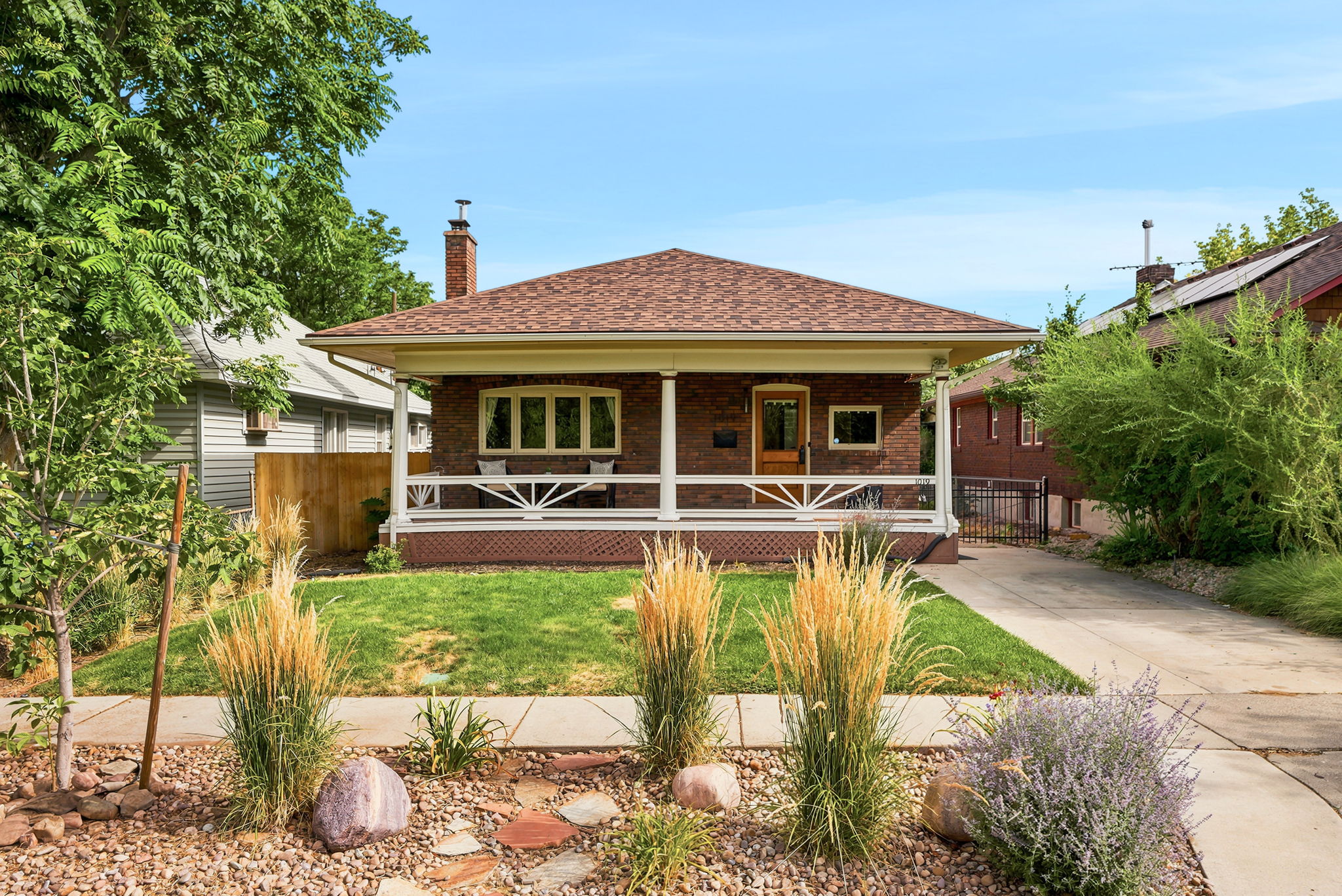 Brick facade residence featuring a covered front porch with white railings, a brown shingle roof, and a mature front yard with decorative plantings