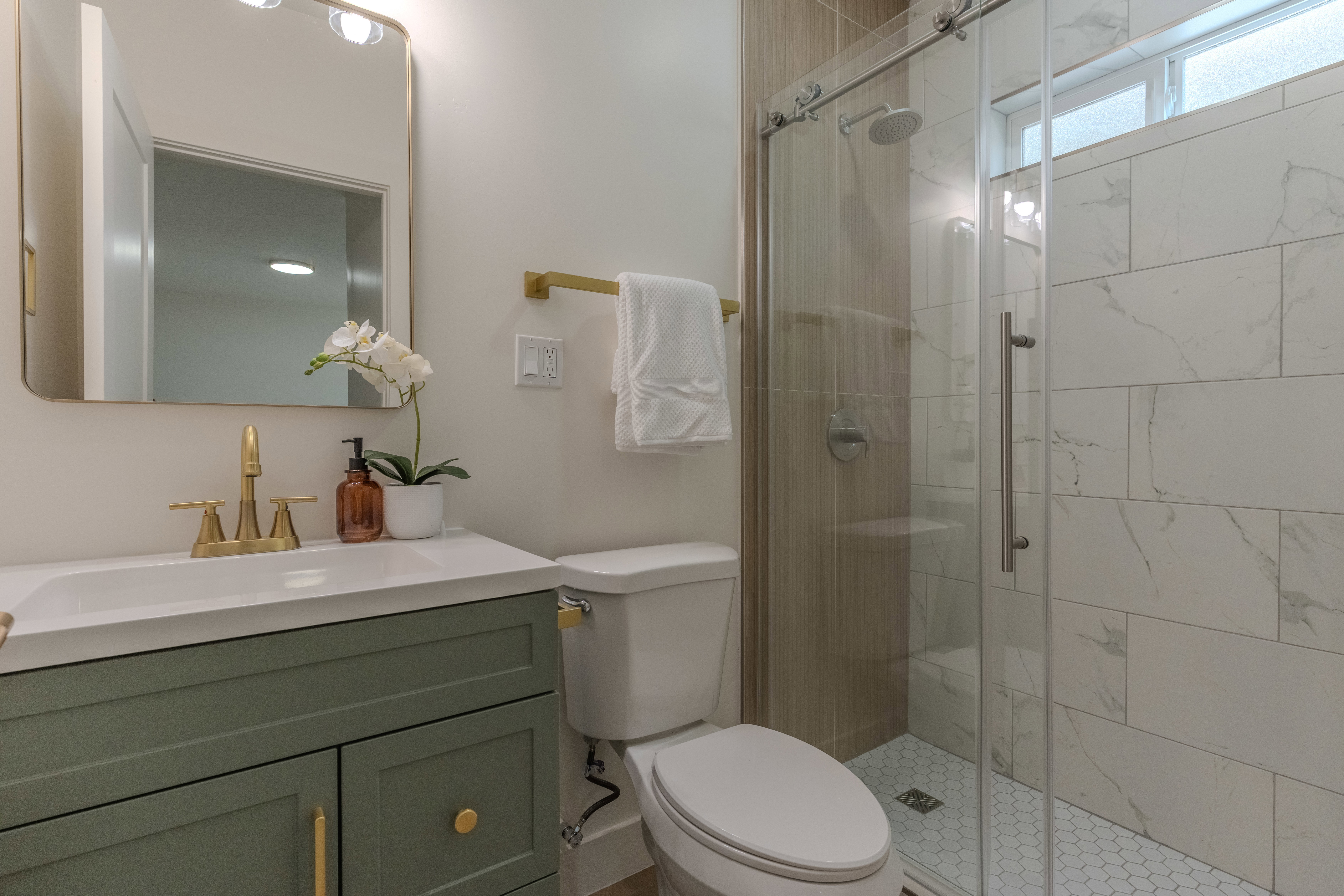 Bathroom featuring a glass-enclosed shower with white hexagonal floor tiling, a white vanity countertop with an integrated sink, a green vanity with gold-tone hardware, and a gold-tone framed mirror