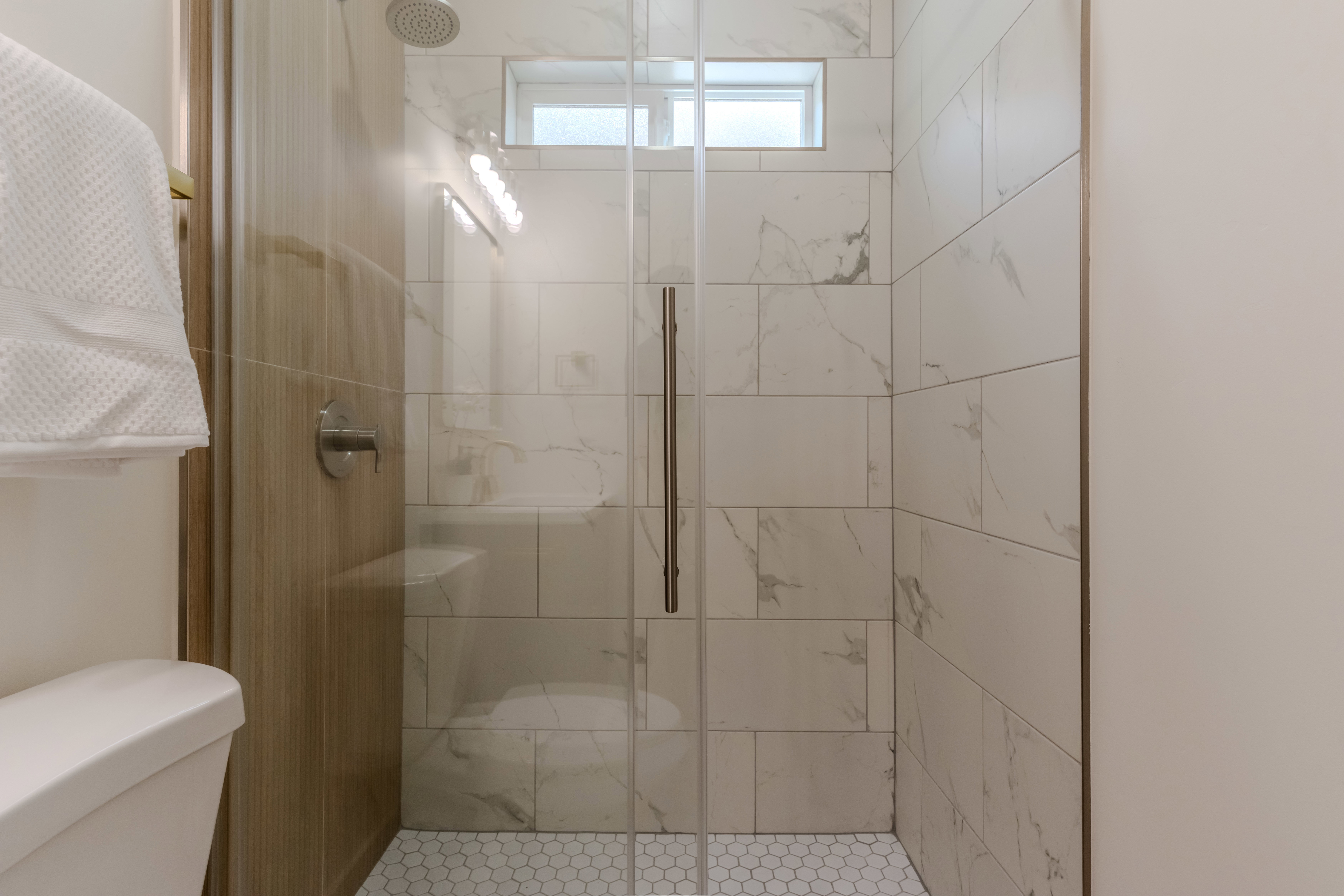 Walk-in shower featuring a clear glass enclosure, large format marble-look tile walls, a rain showerhead, a high window, and a white hexagonal tile floor