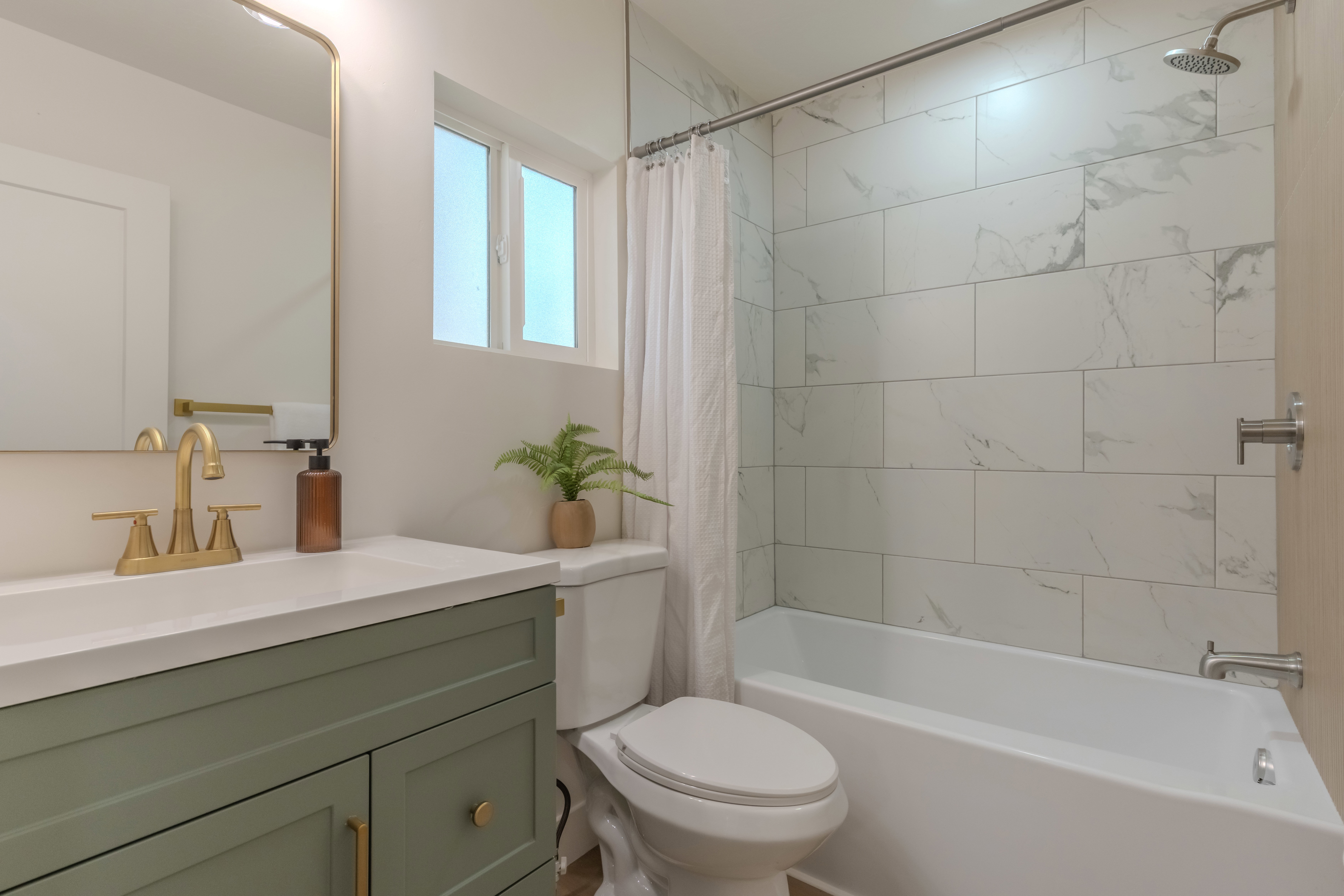 Bathroom featuring a vanity with a light green base and white countertop, gold-toned faucet and hardware, rectangular mirror with integrated lighting, and a white tile shower surround with a rain shower head