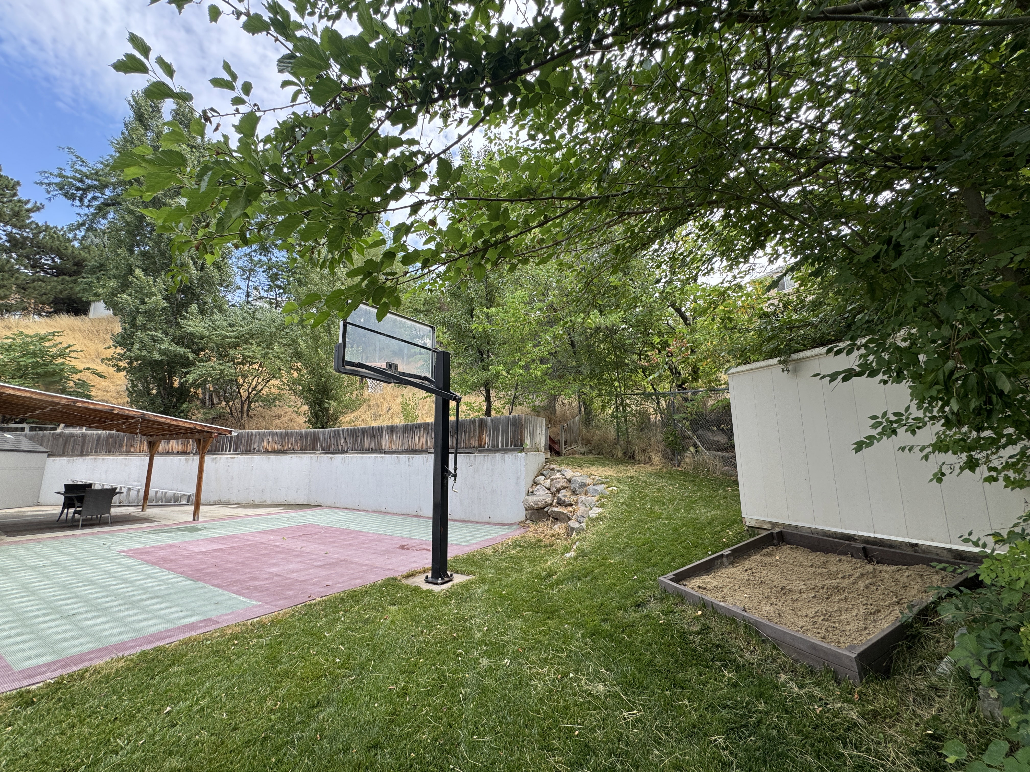 Expansive backyard featuring a multi-sport court with a basketball hoop, a built-in pergola, a functional shed, and a raised garden bed, all surrounded by a wood fence