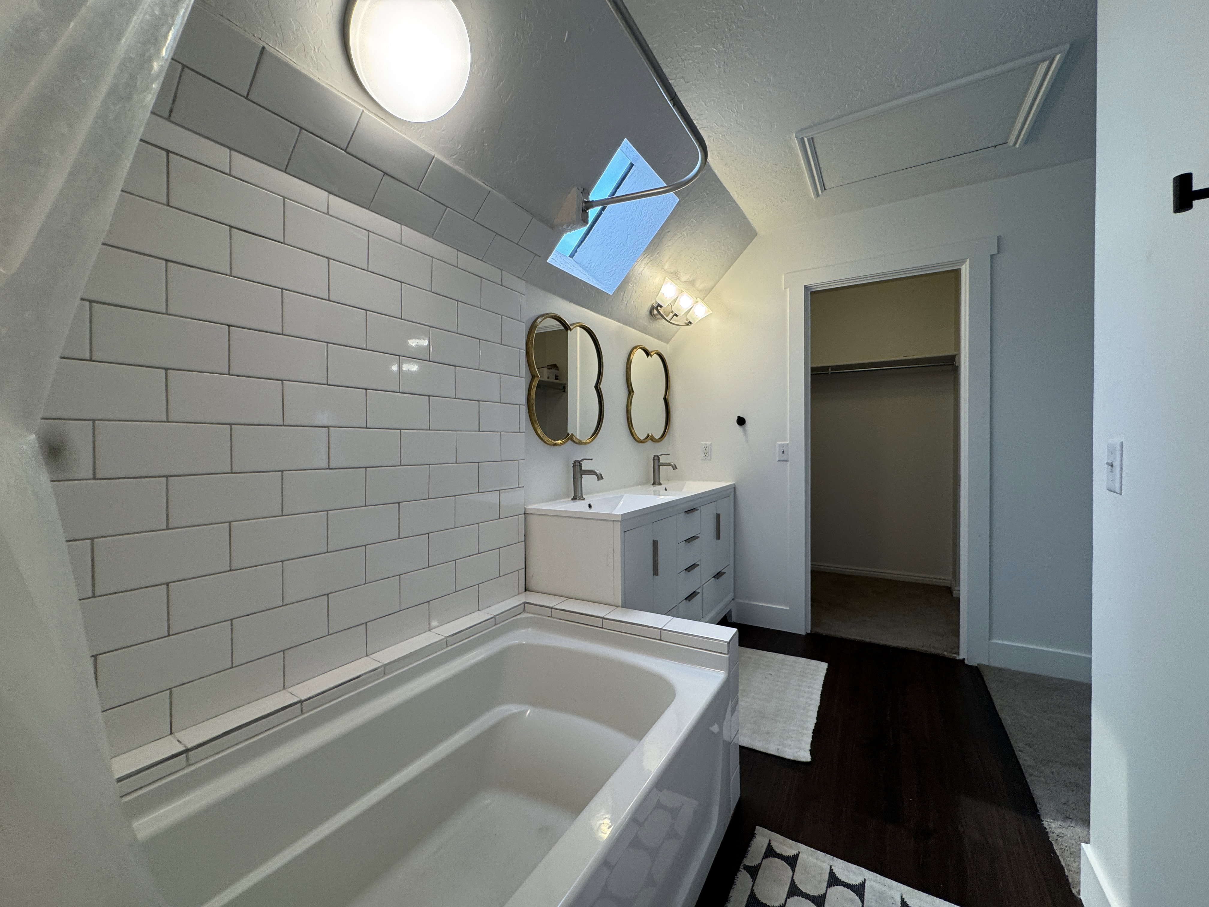 Bathroom featuring a white subway tile surround bathtub, dual sink vanity with modern fixtures, and dark wood-finish flooring