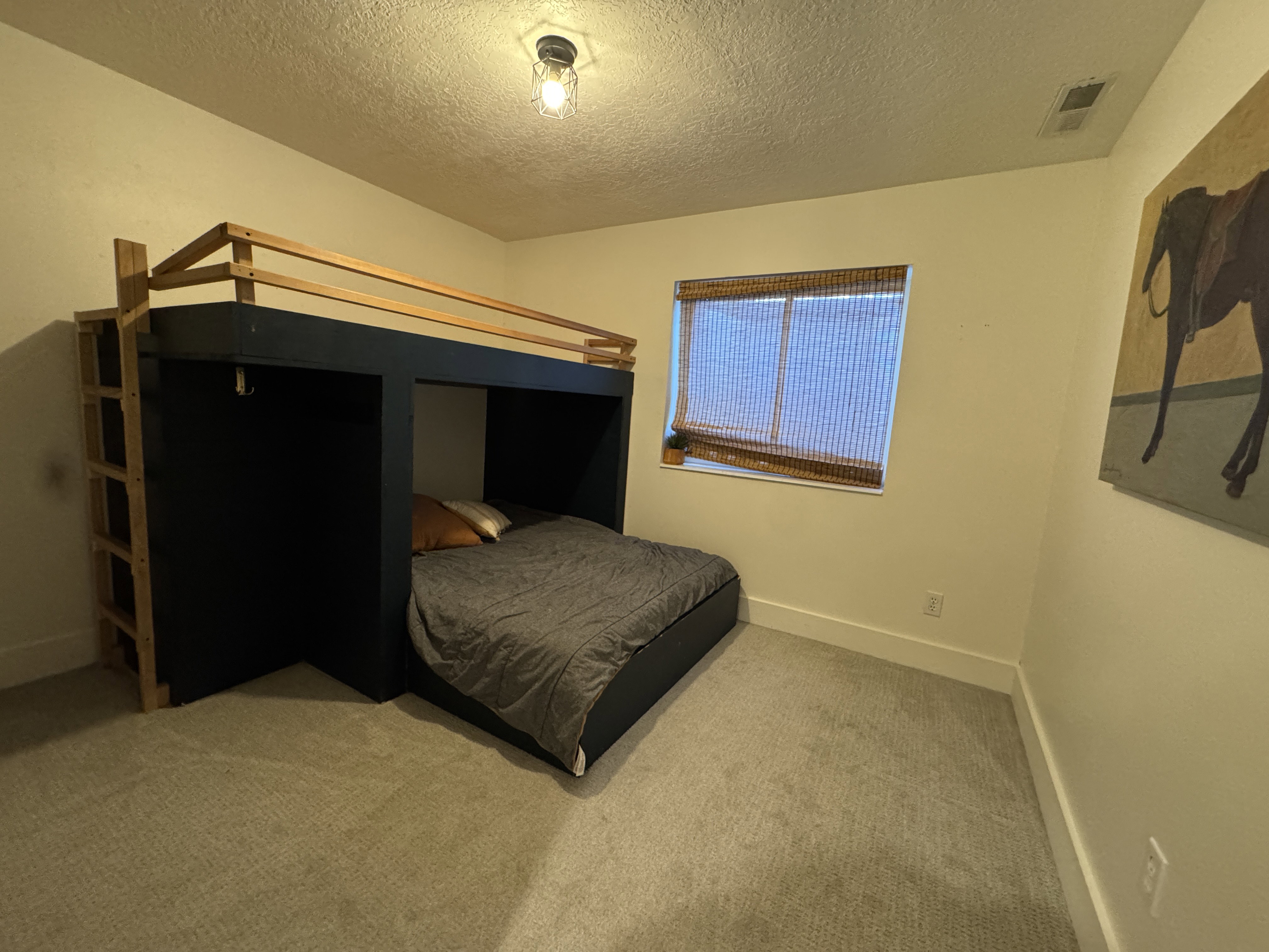 Built-in loft bed with integrated ladder, light-colored walls, recessed window with a woven blind, and gray carpeting
