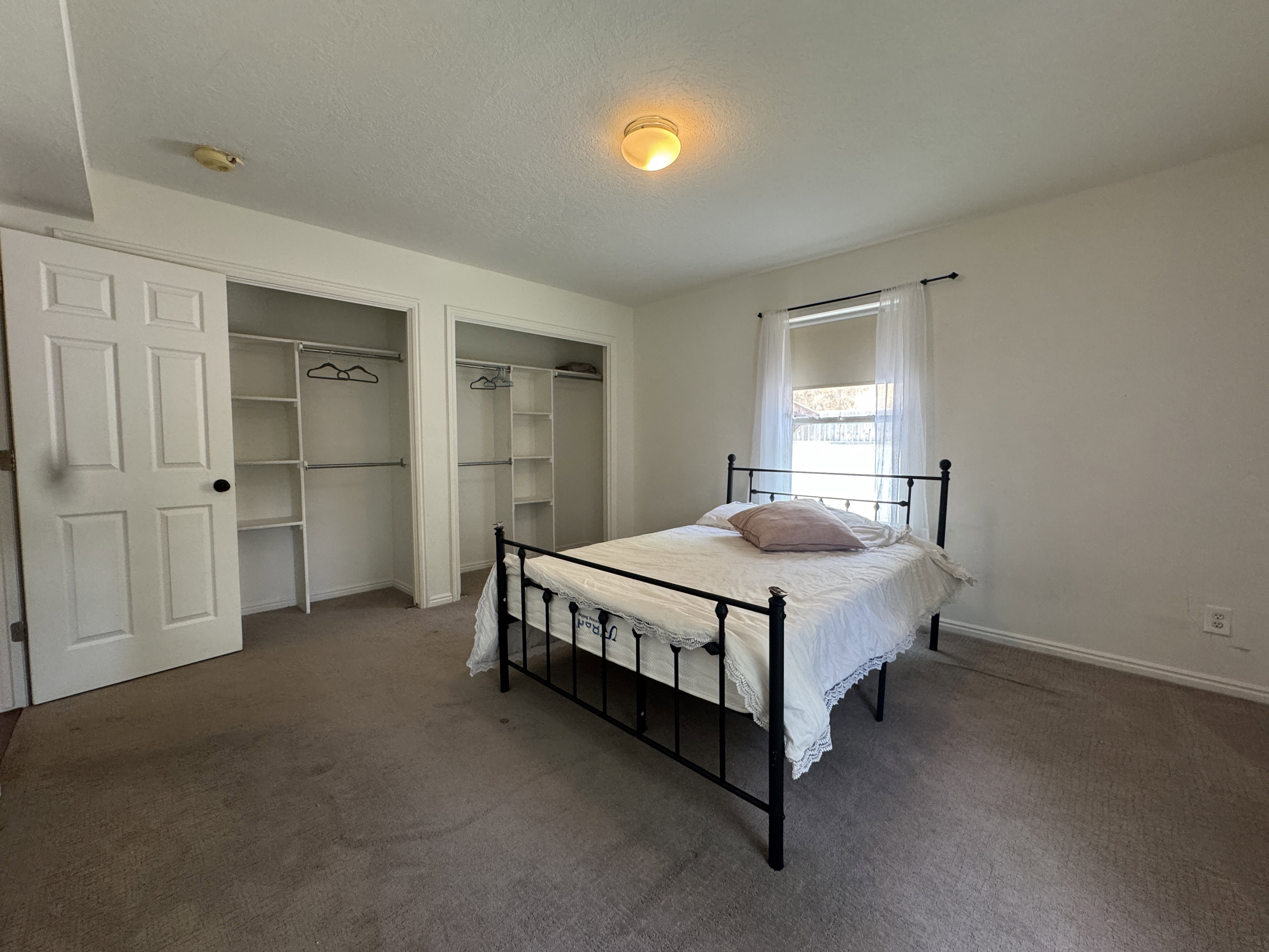 Bedroom featuring two built-in closets with shelving and hanging rods, a window with a roller shade and sheer curtains, carpet flooring, and a flush mount ceiling light