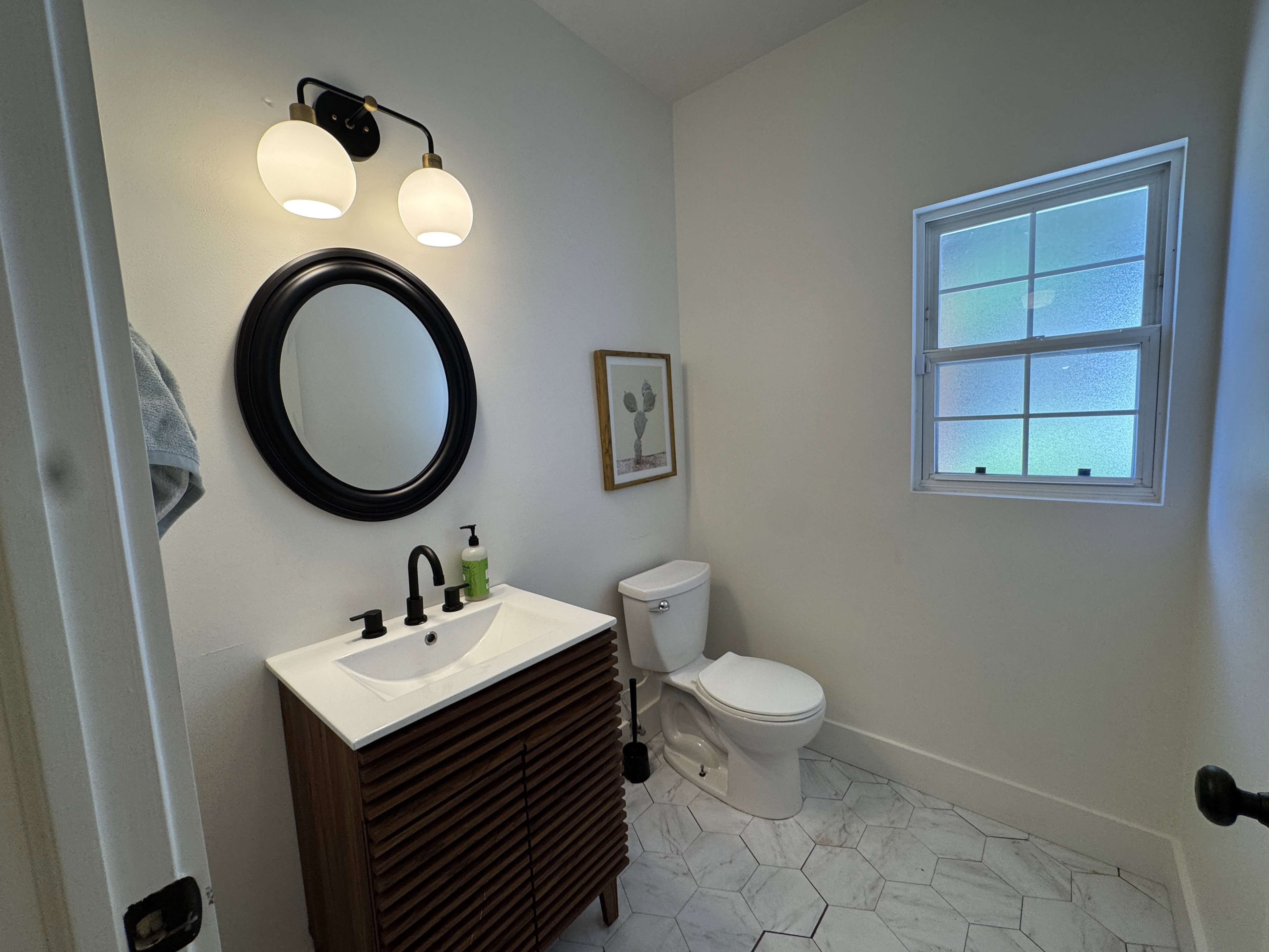 Bathroom featuring a wood-finish vanity with a white countertop and integrated sink, matte black faucet, and a round black-framed mirror