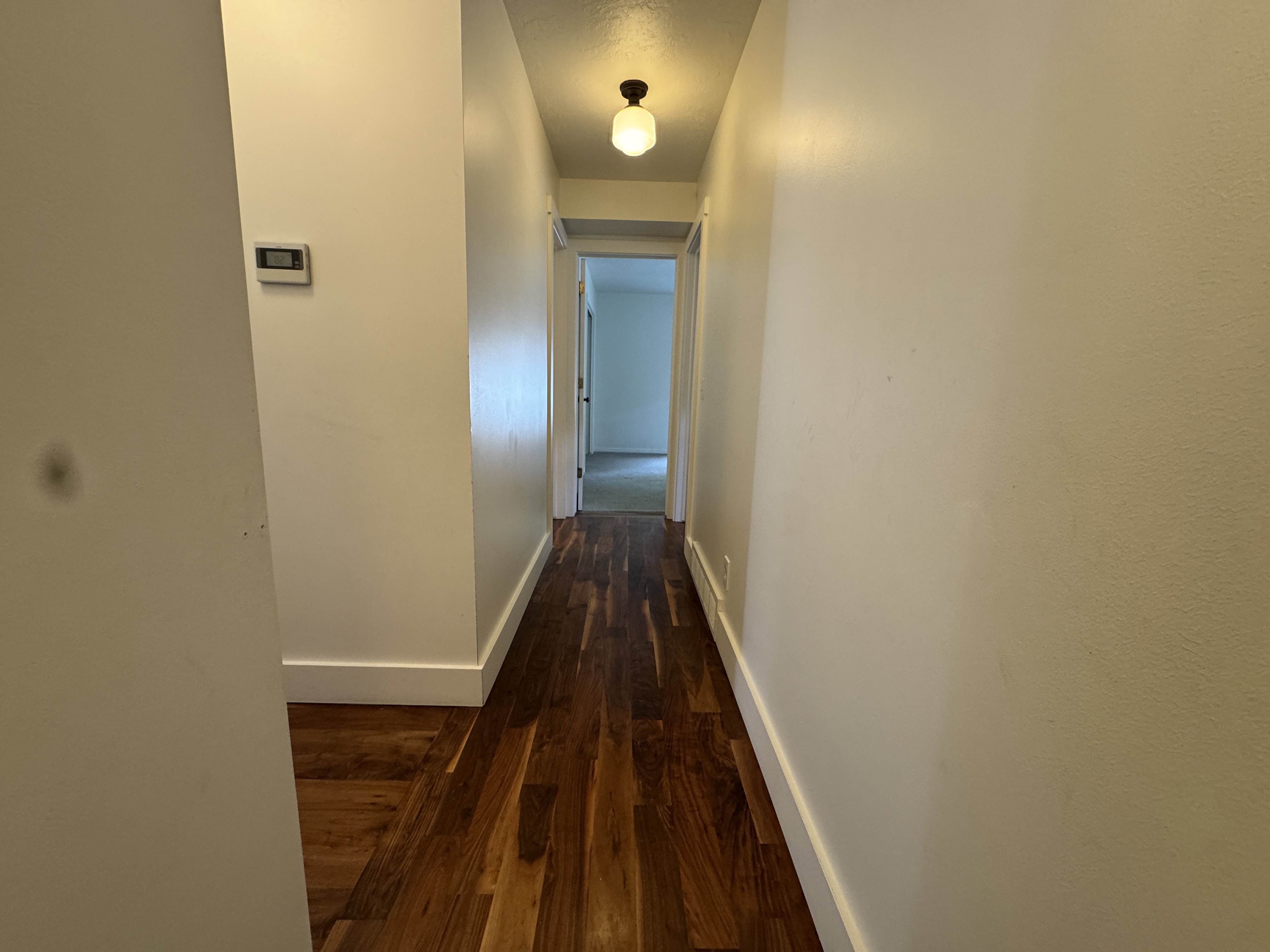 Hallway featuring wood-finish flooring, white baseboards, a ceiling-mounted light fixture, and a wall-mounted thermostat