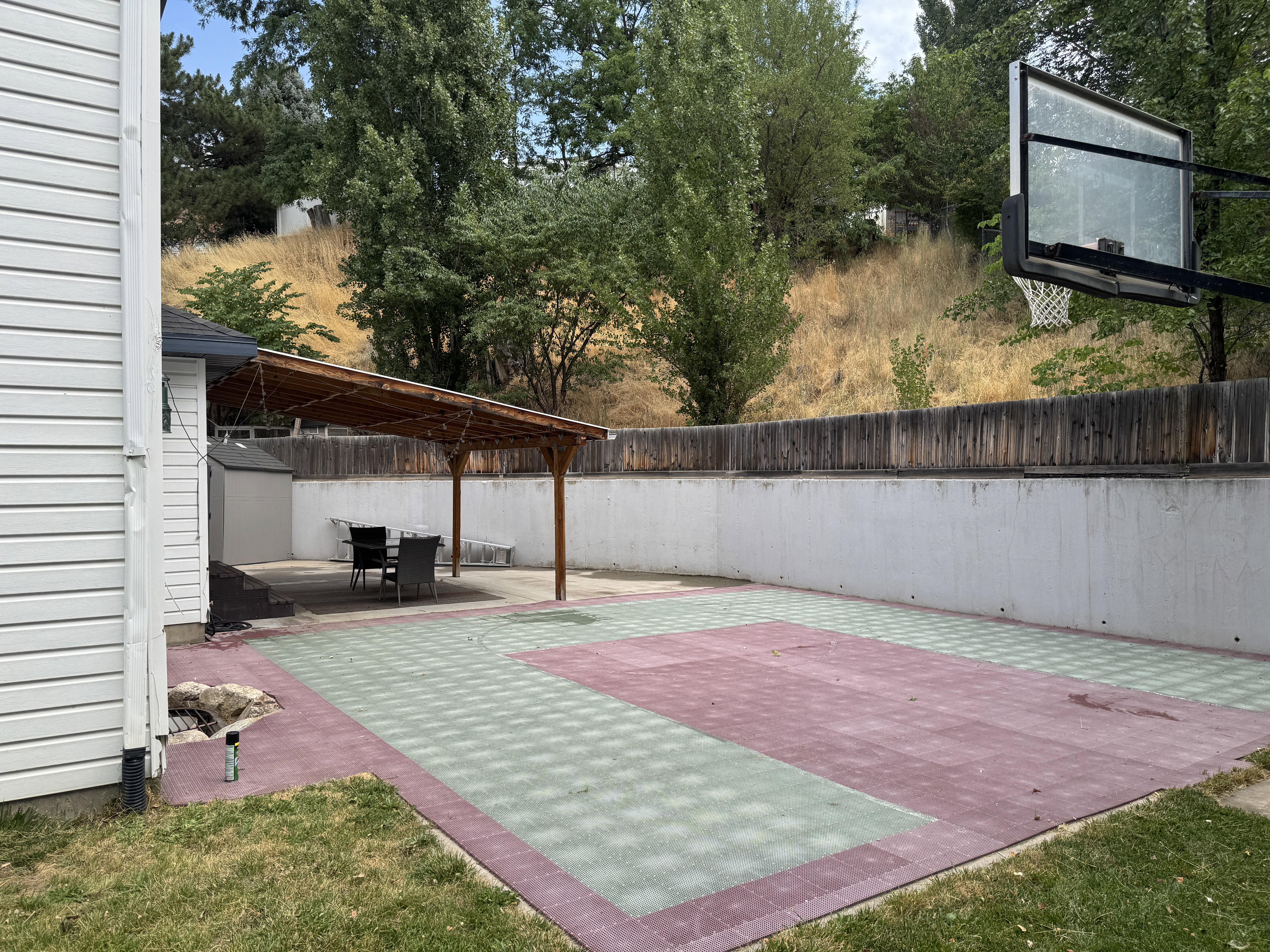 Expansive backyard featuring a multi-sport court with red and green surfacing, a concrete retaining wall topped with wood fencing, and a built-in basketball hoop