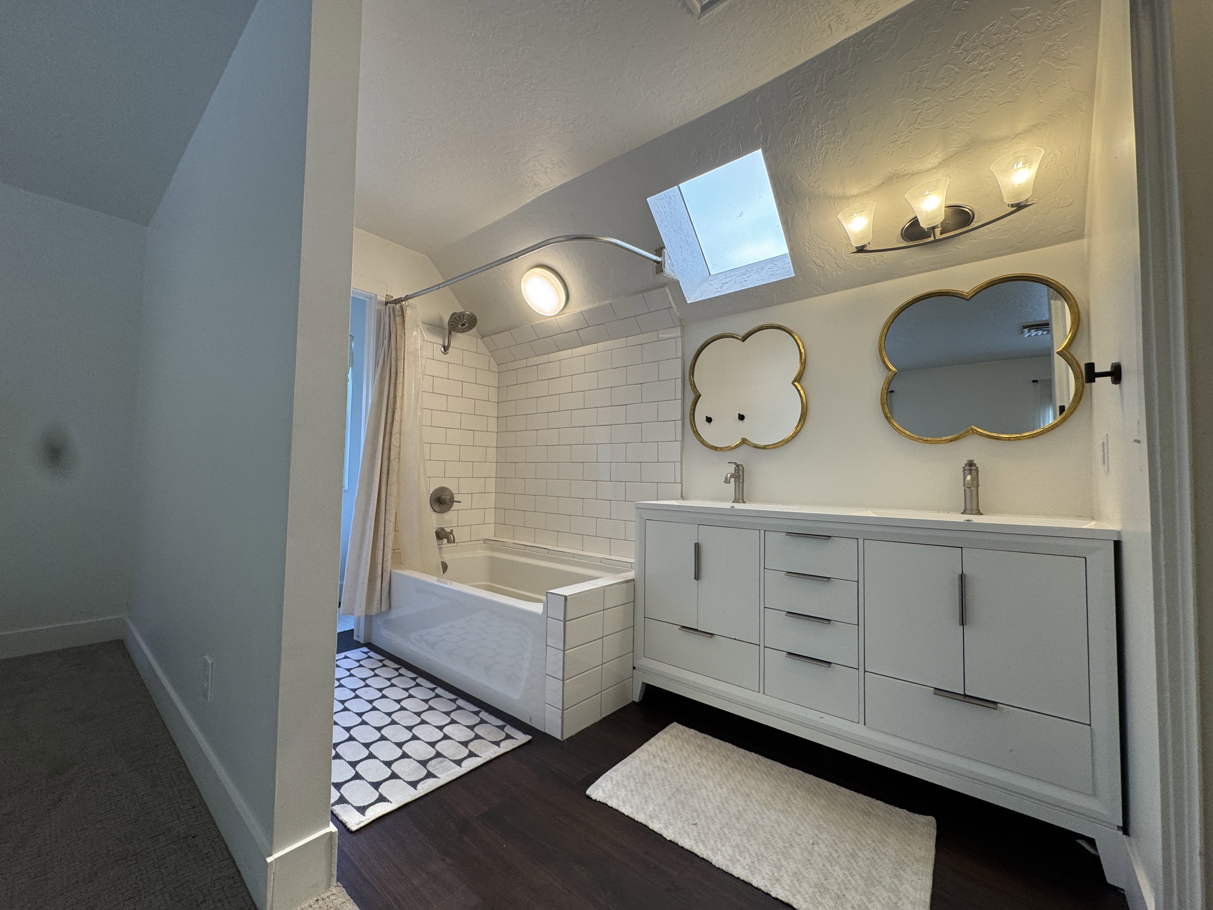 Bathroom featuring a double vanity with ample storage and undermount sinks, two decorative wall mirrors, a skylight, and dark wood-finish flooring