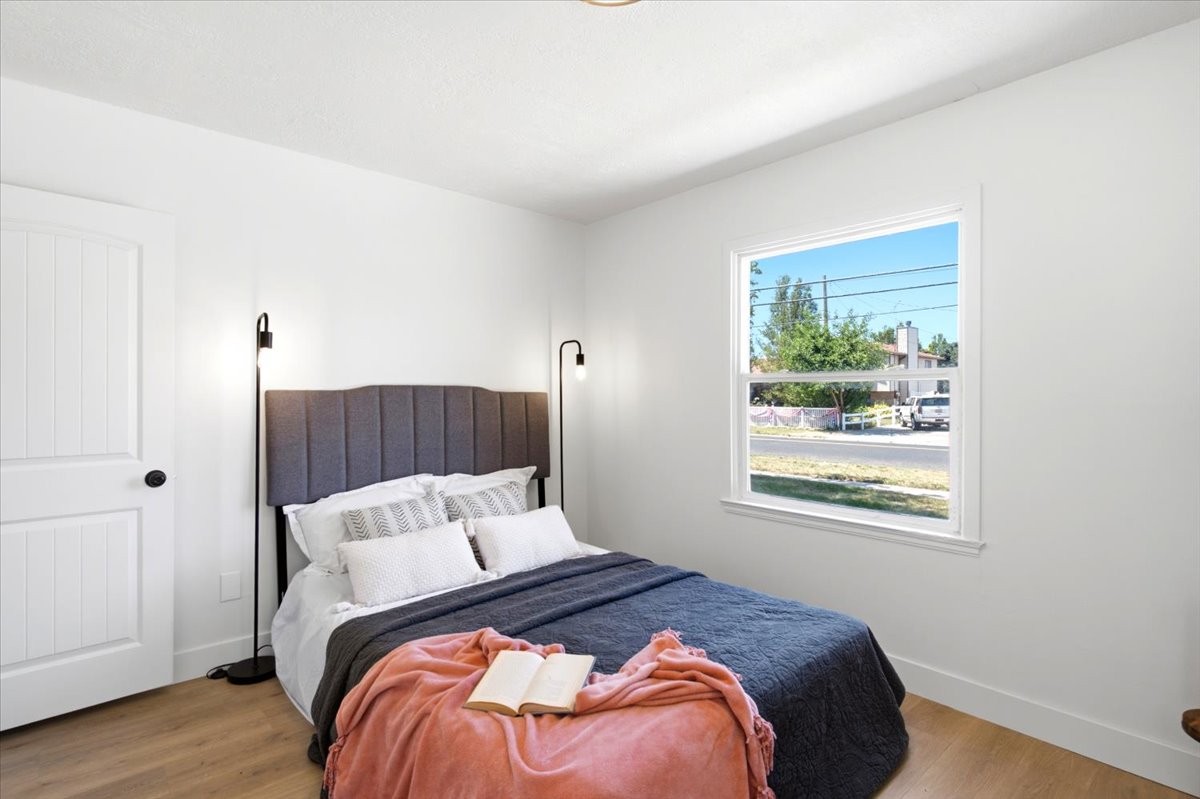 Bedroom featuring wood-finish flooring, a white interior door with matte black hardware, white baseboards, and a large window with white trim
