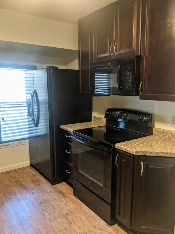 Kitchen featuring dark wood-finish cabinetry with silver hardware, granite-look countertops, black oven with stovetop, black microwave, and black refrigerator