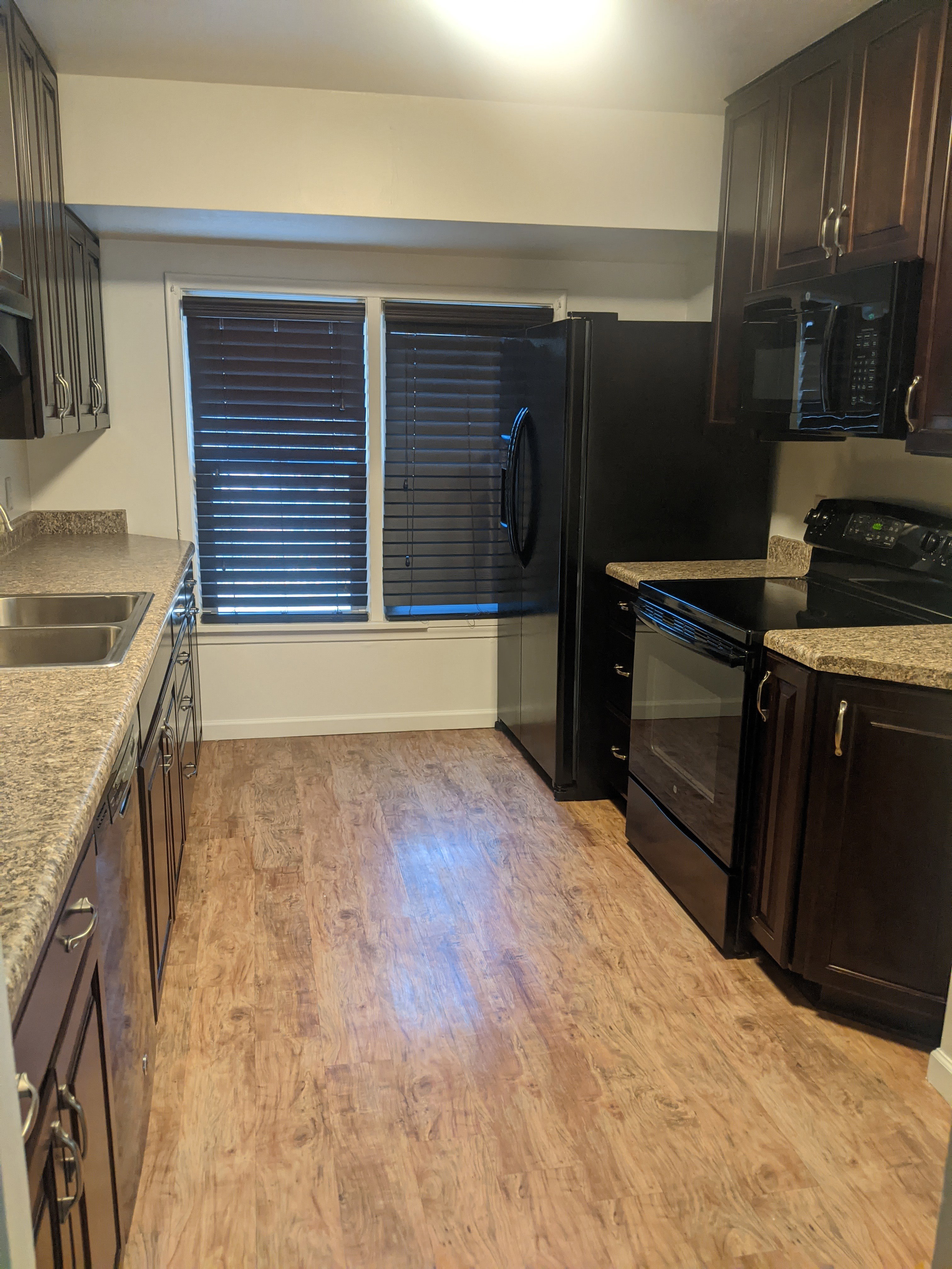 Galley kitchen featuring wood-finish flooring, dark wood cabinetry, granite-style countertops, and a full suite of black appliances