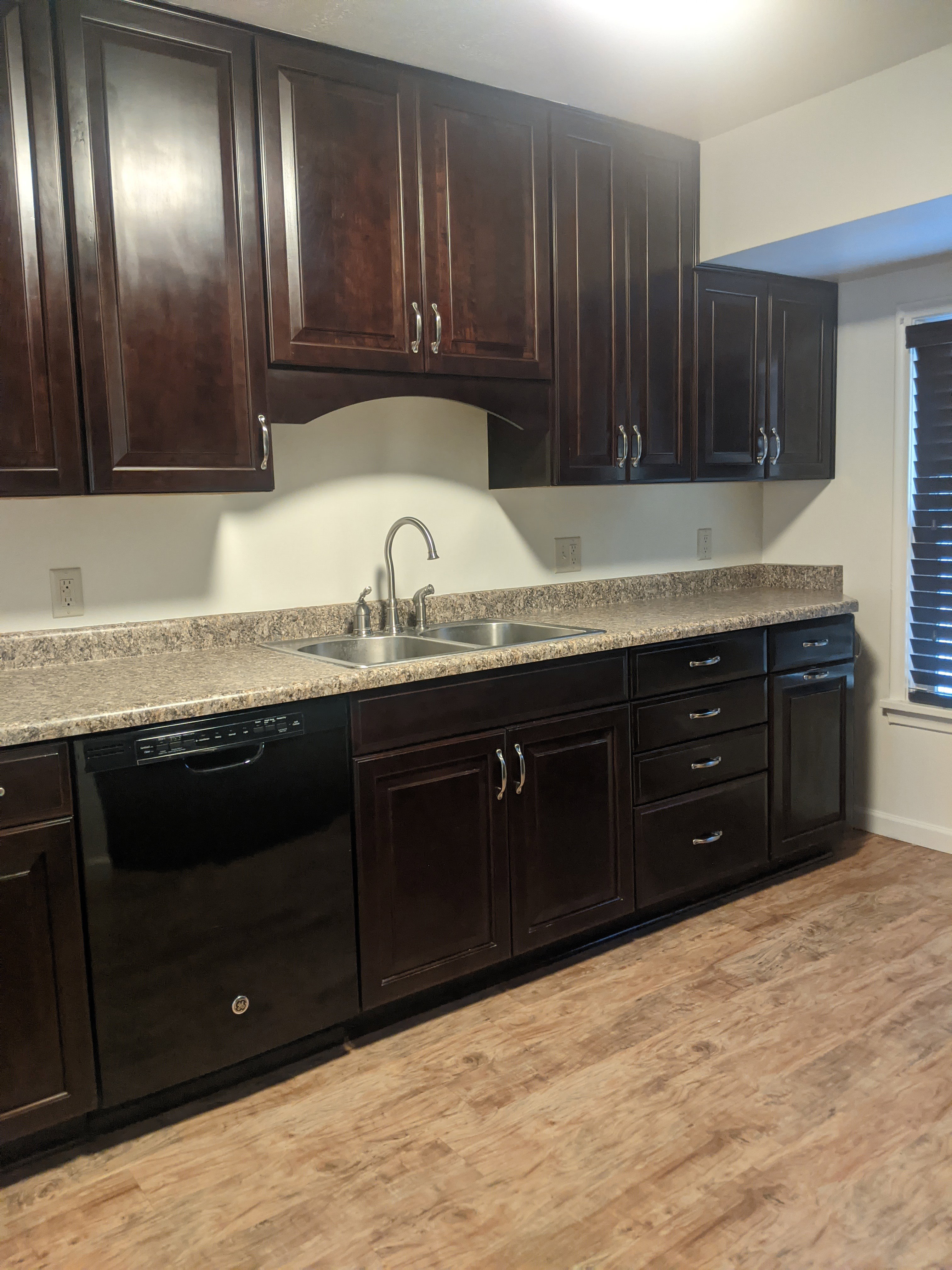 Kitchen featuring dark wood-finish cabinetry with silver-tone hardware, a double basin stainless steel sink, speckled countertops, and wood-finish flooring