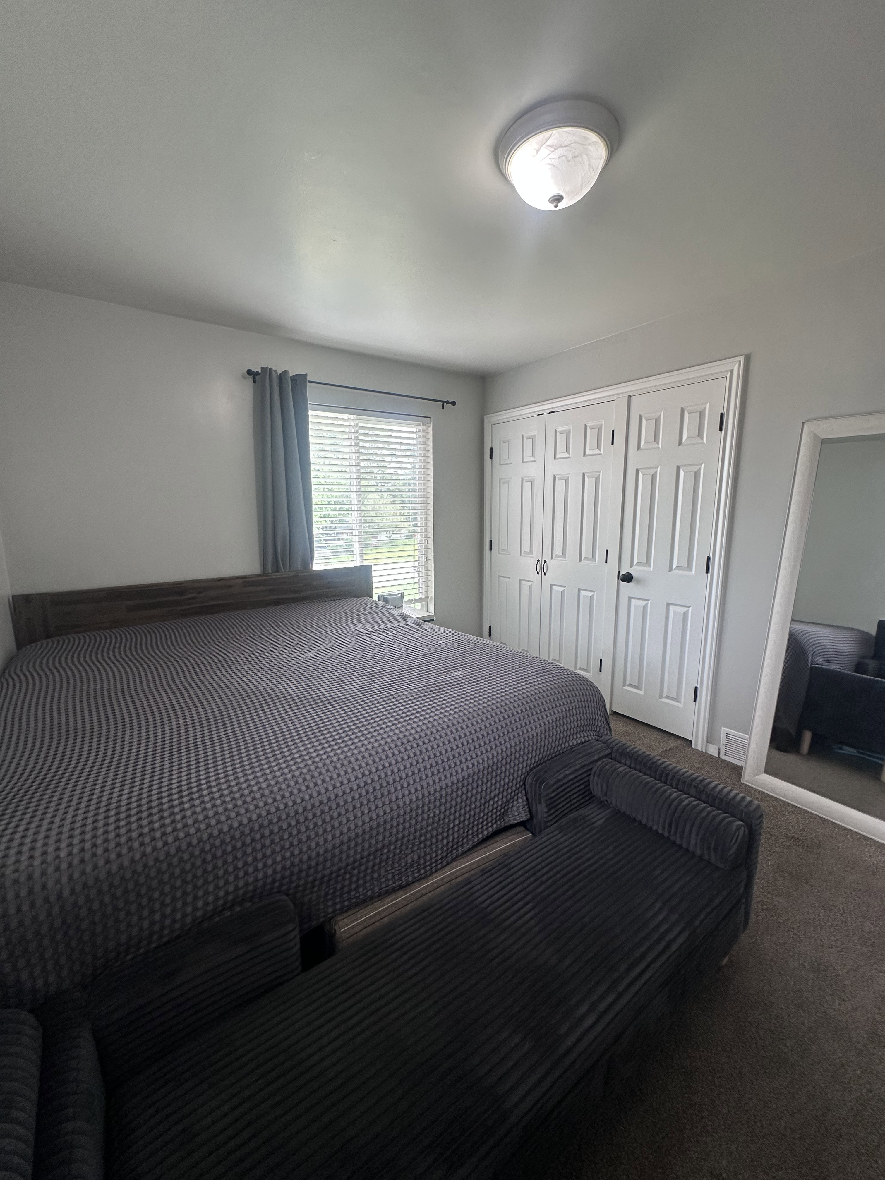 Bedroom featuring light gray walls, a window with blinds and blue curtains, white bi-fold closet doors with black hardware, and a ceiling-mounted light fixture
