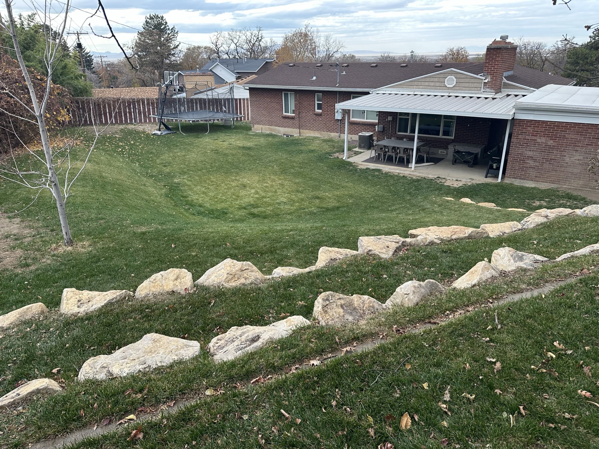 Expansive terraced backyard featuring a manicured lawn, stone retaining walls, privacy fencing, a covered patio, and a brick exterior