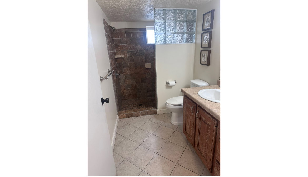 Bathroom featuring a walk-in shower with brown tile surround, glass block window, wood-finish vanity with an integrated sink, and neutral-toned floor tiles
