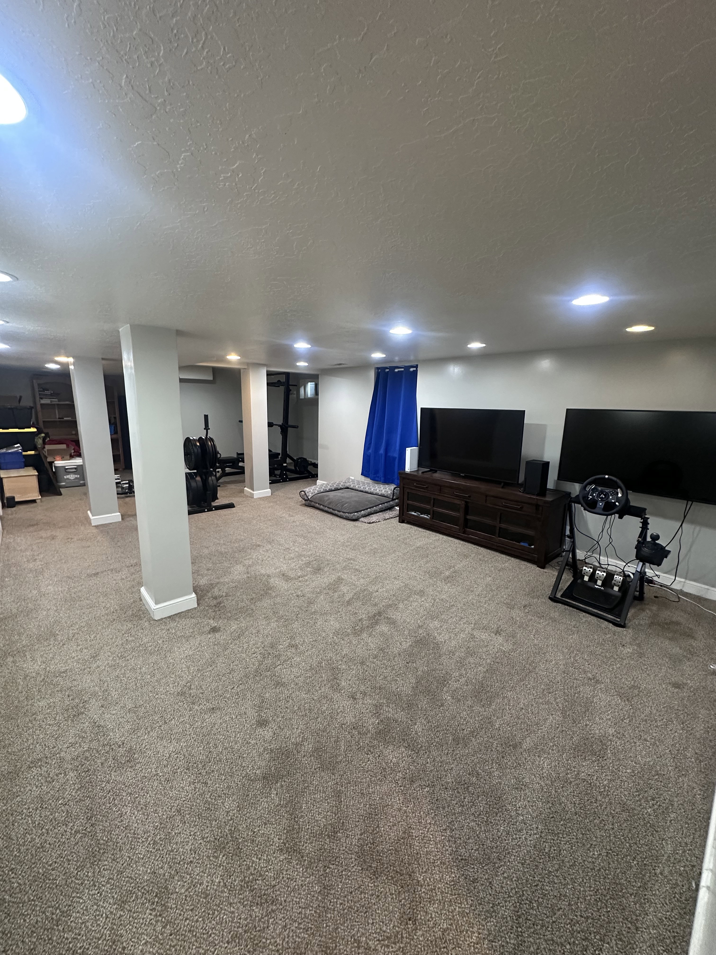 Spacious room featuring neutral carpeting, recessed lighting, support pillars with base molding, a wall-mounted AC unit, and a window with blue drapes