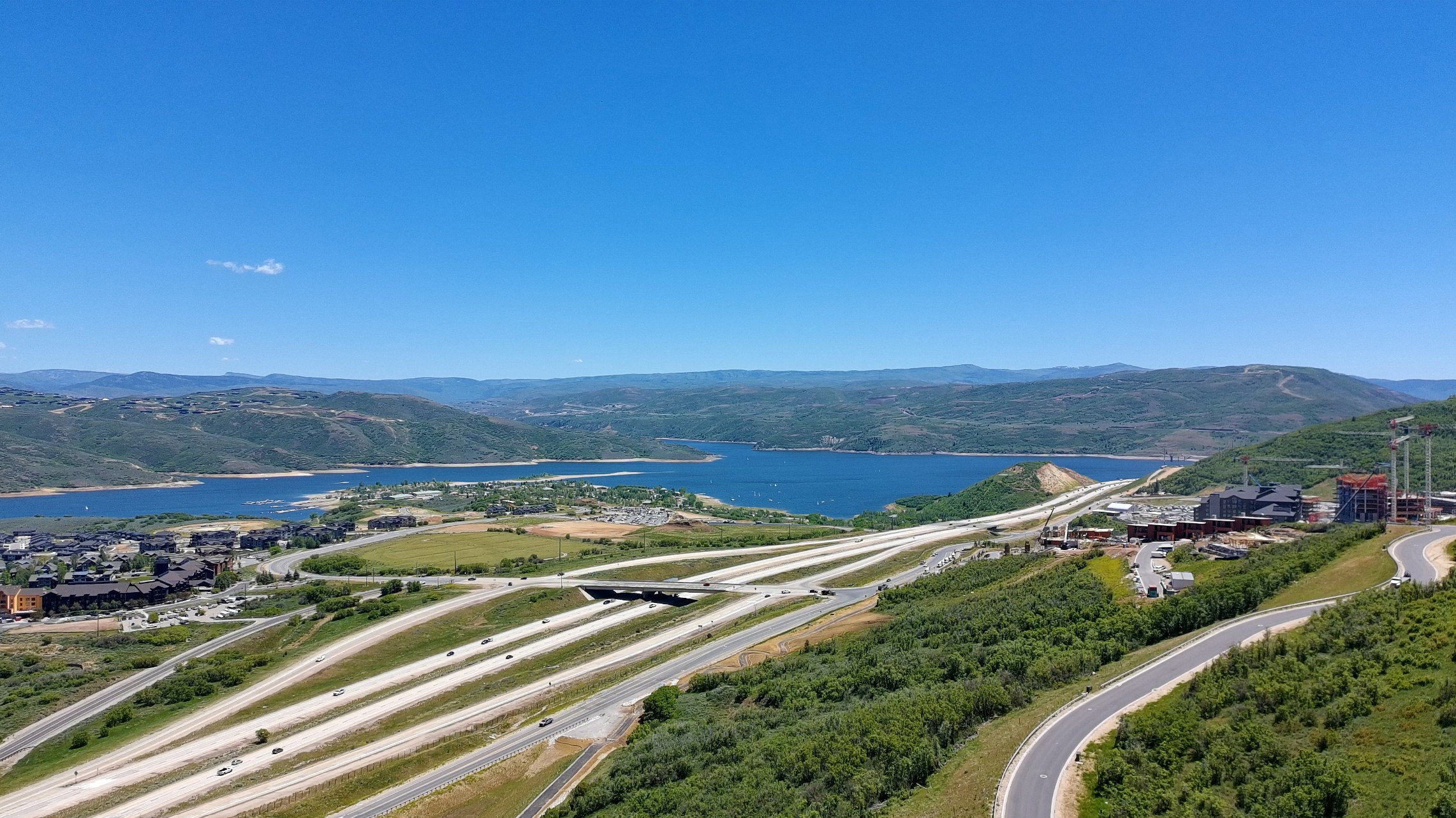 Expansive mountain landscape featuring a large body of water, distant hills, a multi-lane highway, and scattered buildings