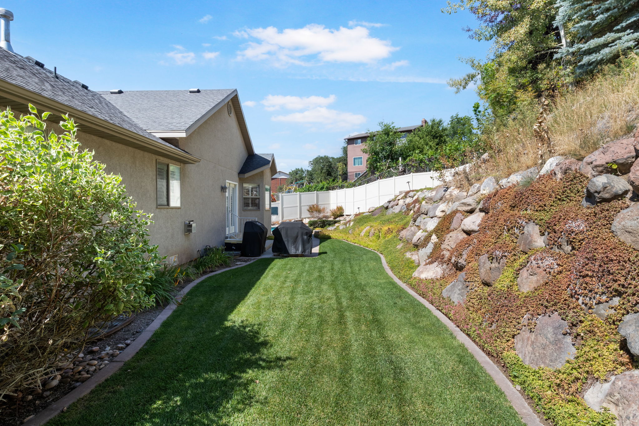 Spacious backyard featuring a manicured lawn, a rock retaining wall with ground cover, and a stucco exterior