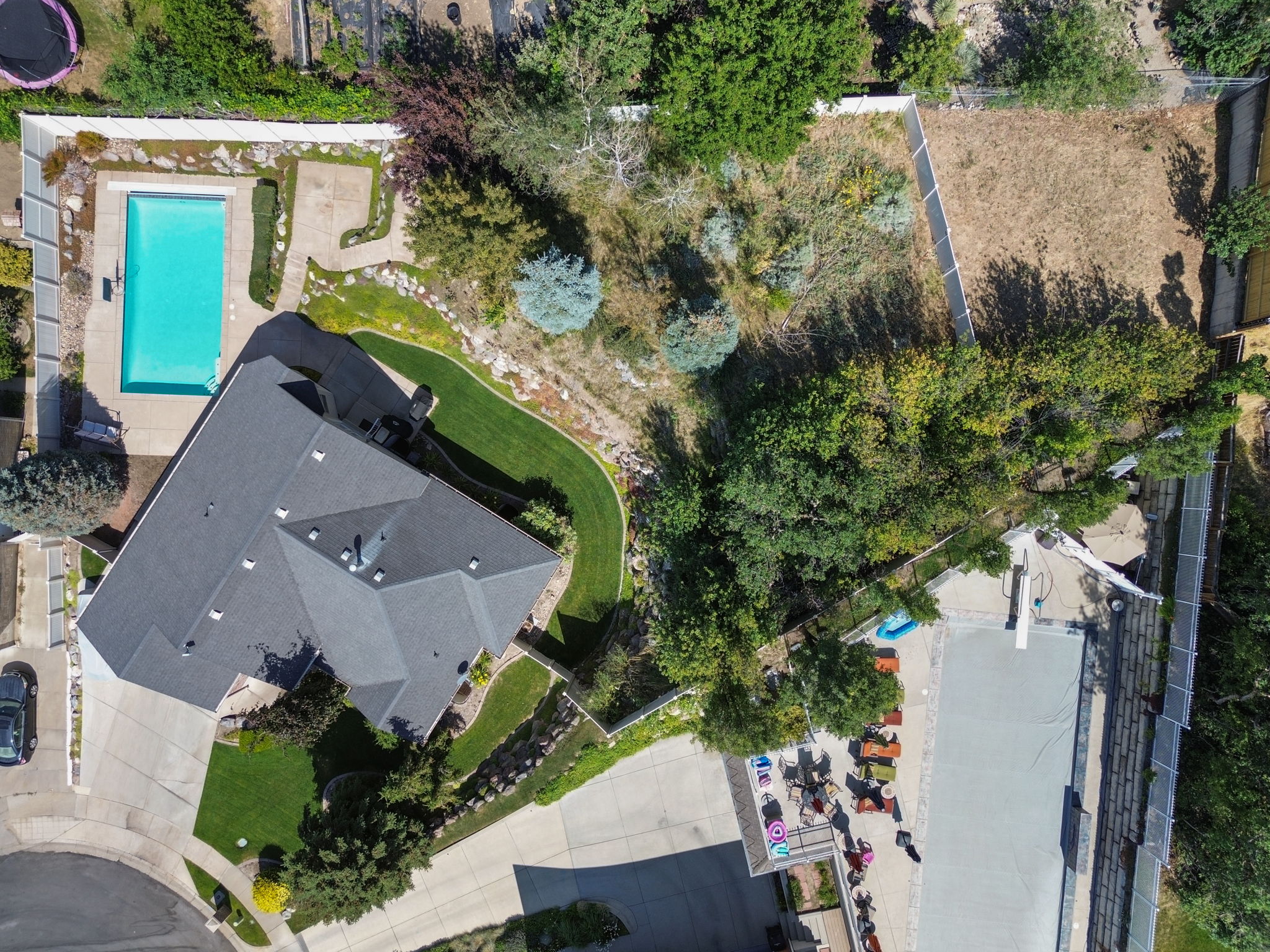 Expansive property featuring a dark-toned roof, multiple paved areas, a built-in swimming pool, mature perimeter landscaping, and an additional covered pool area