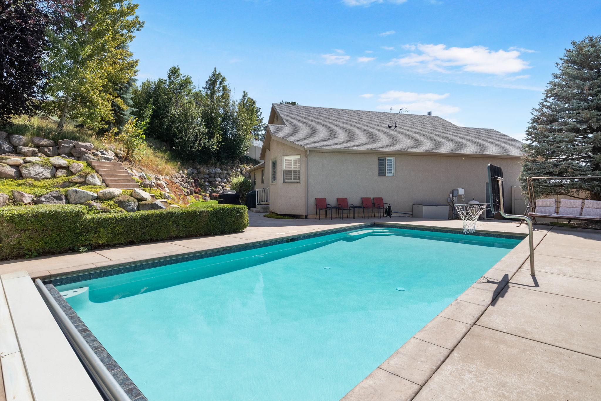 Outdoor swimming pool with concrete surround, bordered by a manicured hedge and a rock retaining wall
