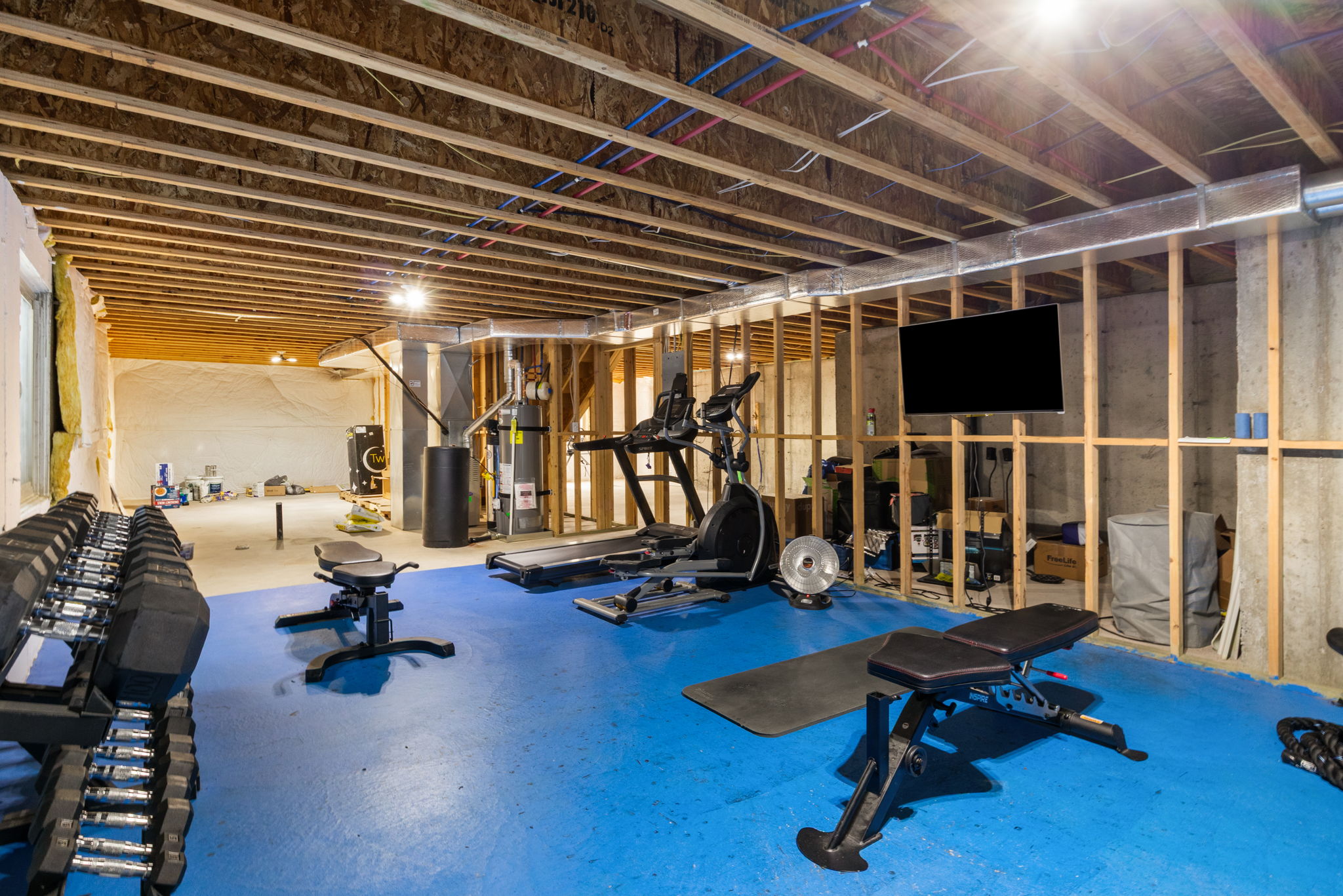 Unfinished basement featuring exposed ceiling joists and wall framing, concrete flooring with a distinct blue section, visible utility pipes, and recessed lighting
