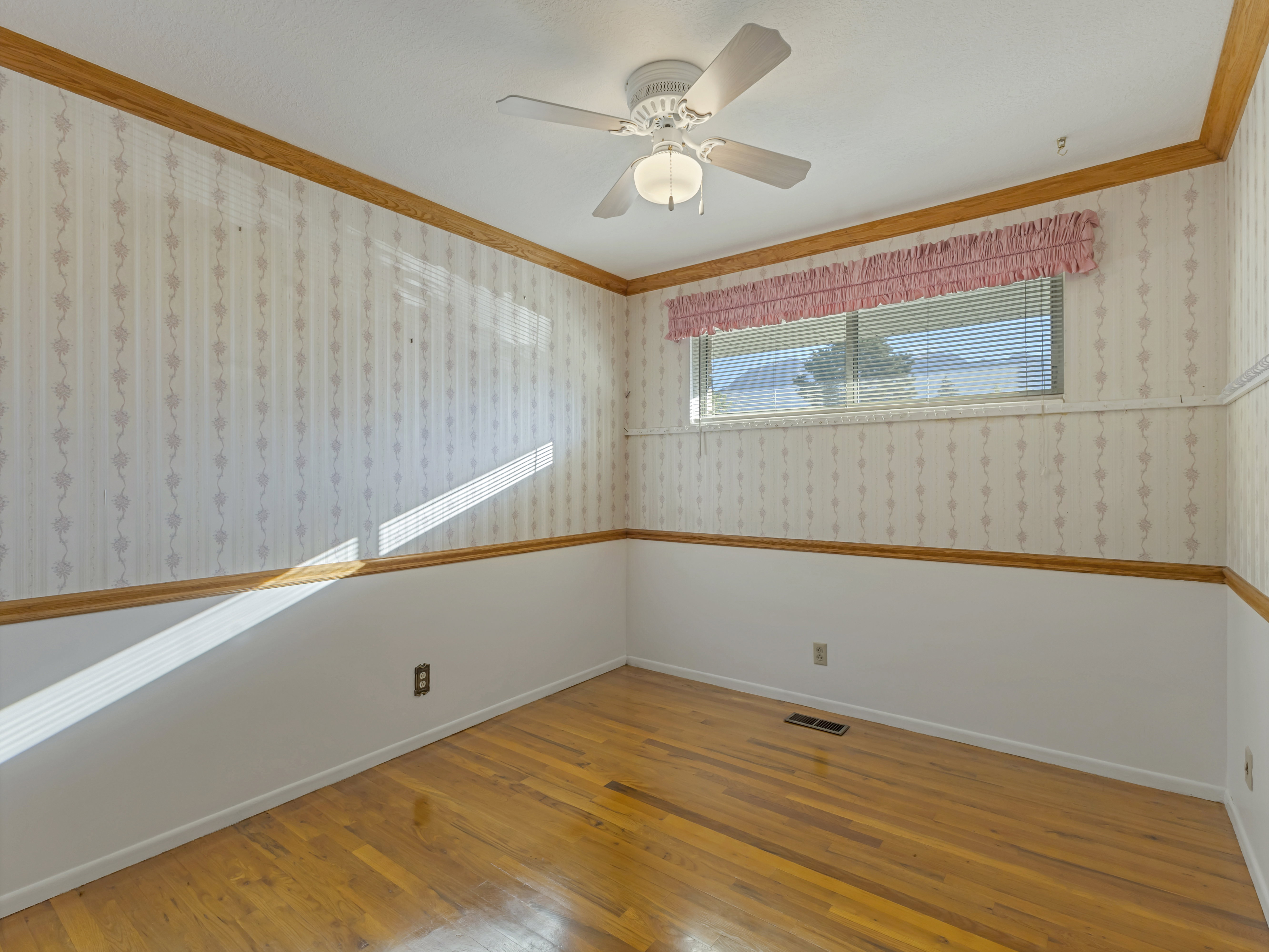 Room featuring wood-finish flooring, white ceiling fan with light fixture, and a window with horizontal blinds and a decorative valance