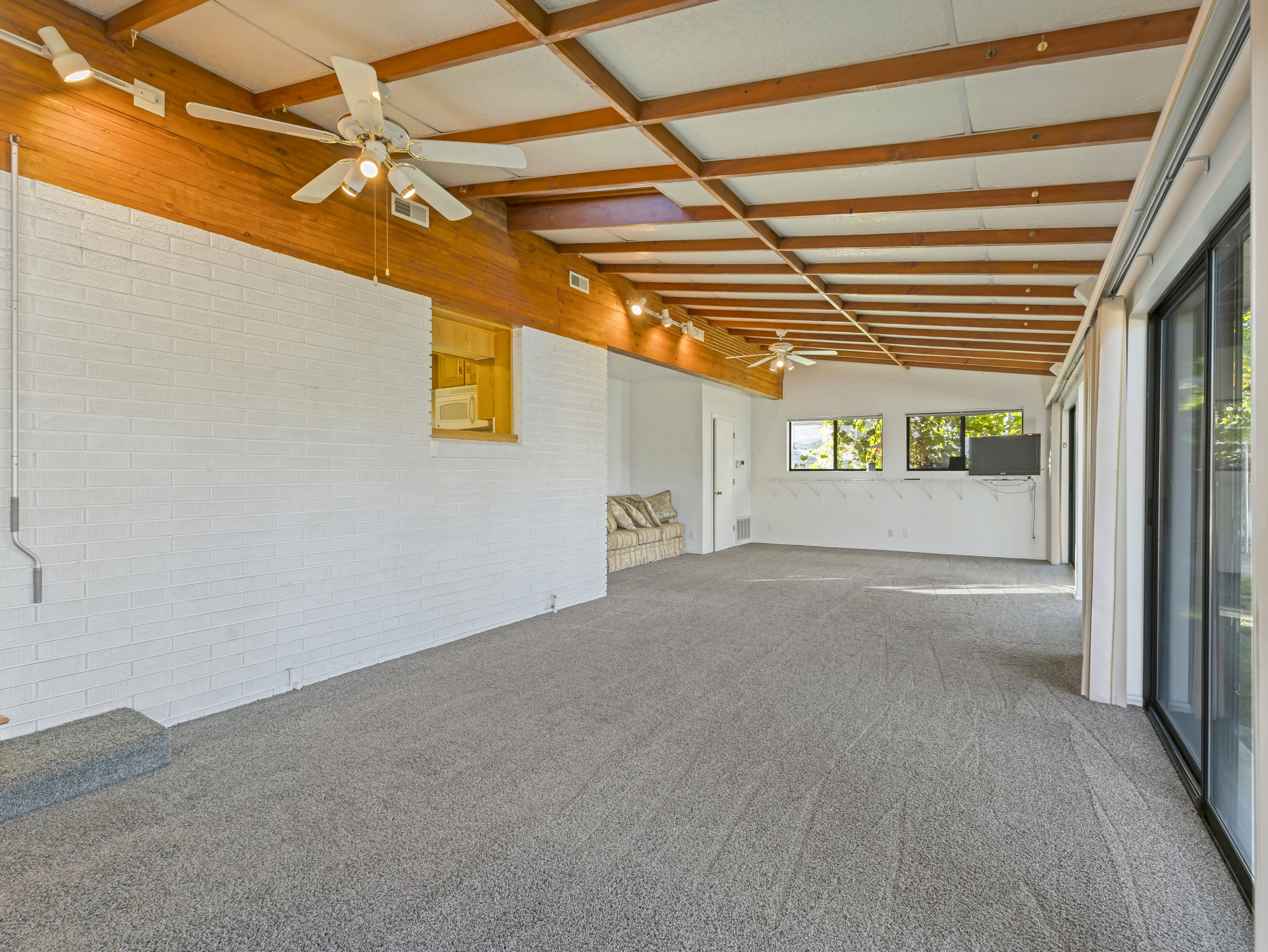 Spacious room with exposed wood beam ceiling, white painted brick wall, gray carpet flooring, and multiple ceiling fans