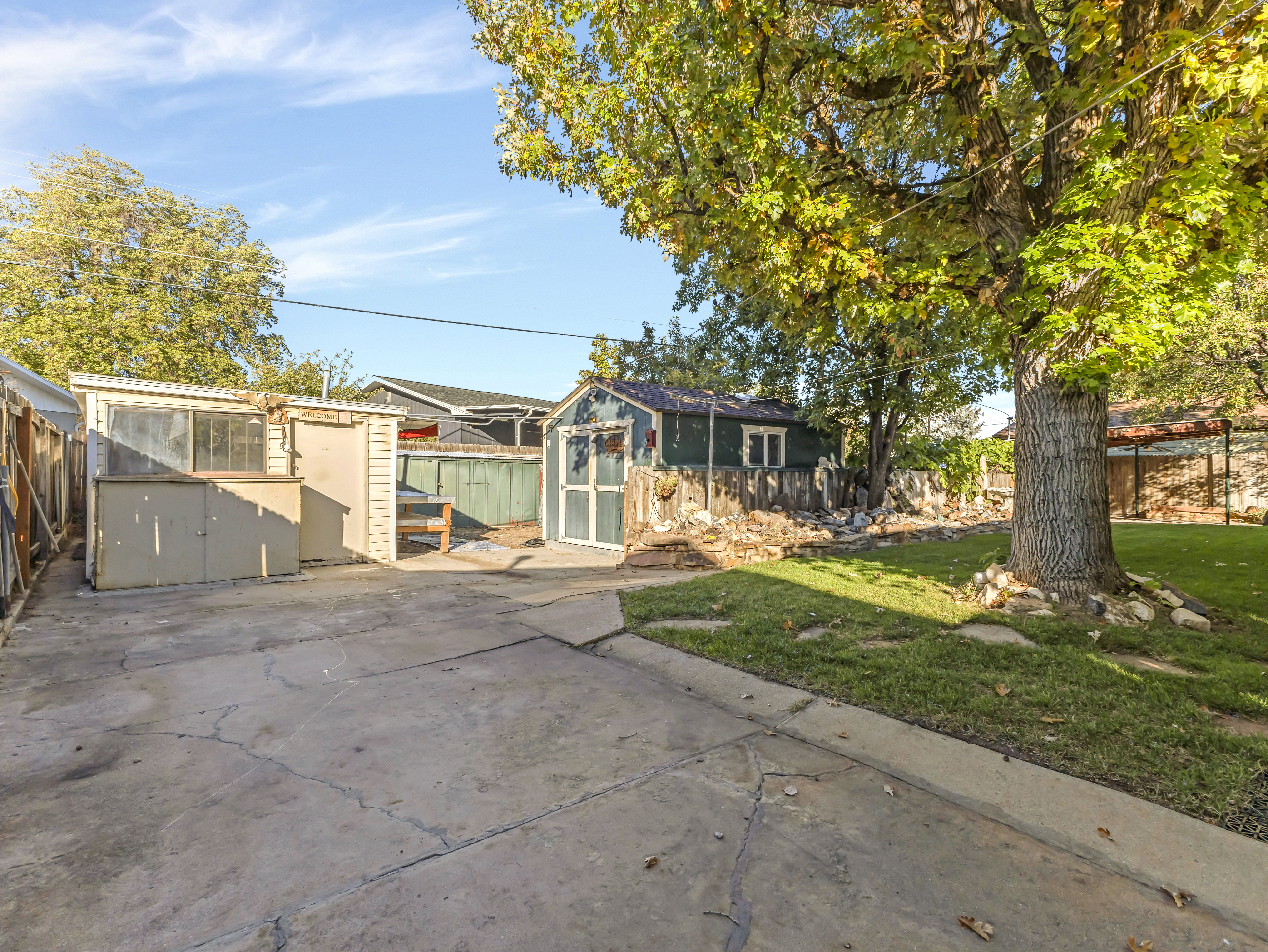 Concrete patio area featuring two storage sheds, a mature deciduous tree, and a grass lawn