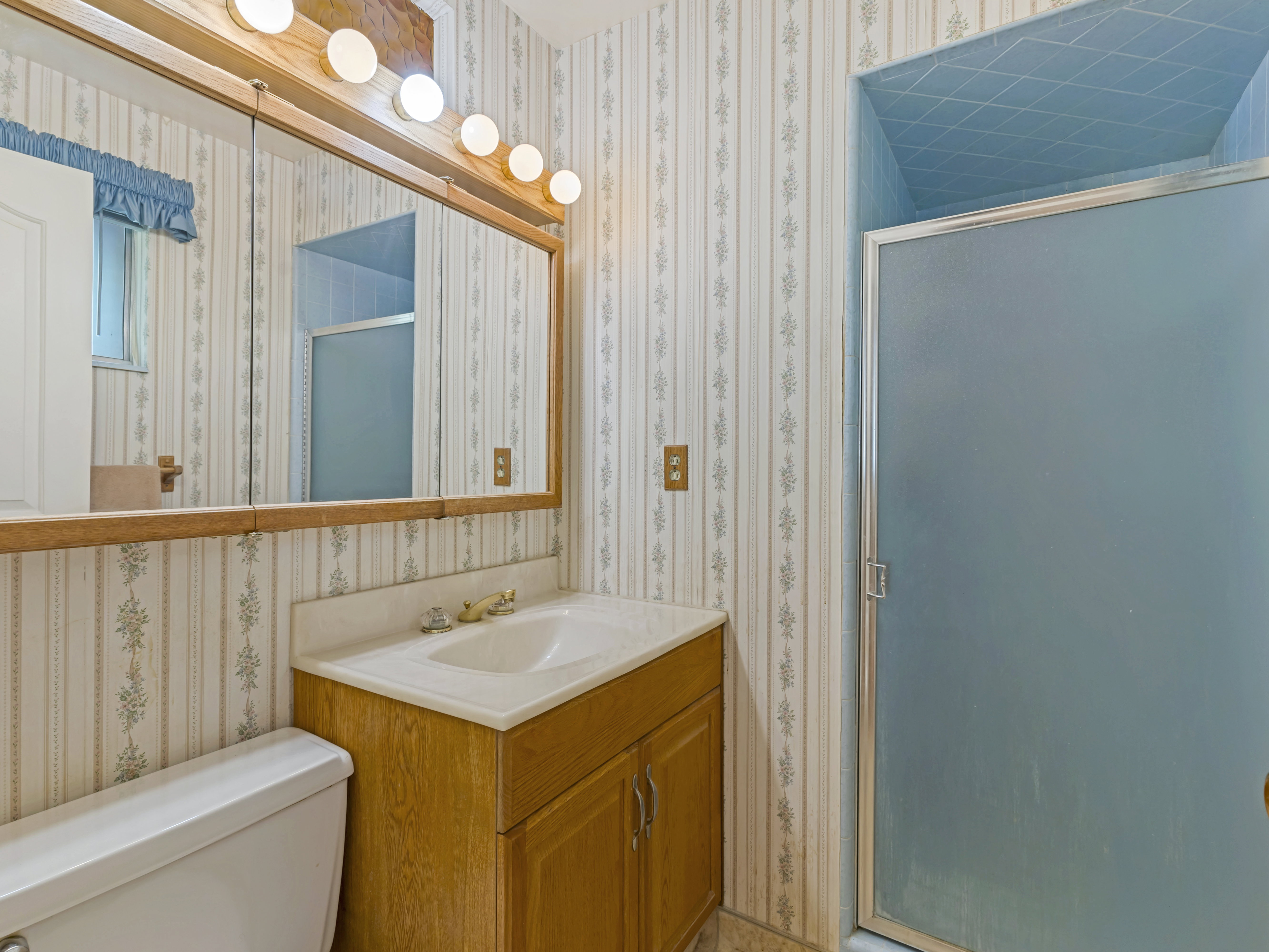 Bathroom featuring a vanity with an integrated sink, a wall-mounted mirror with integrated lighting, a toilet, and a stand-up shower with a frosted glass door