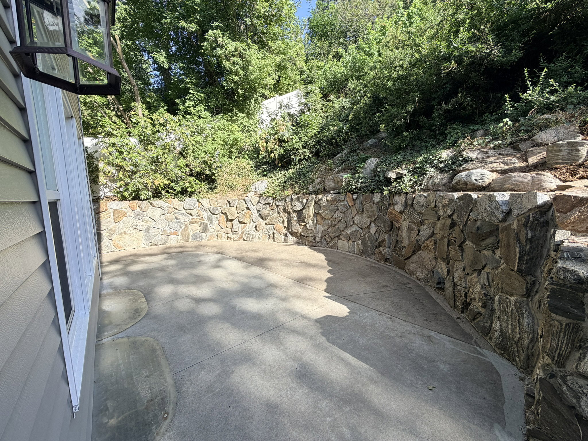 Spacious concrete patio featuring a curved stone retaining wall and surrounding mature foliage