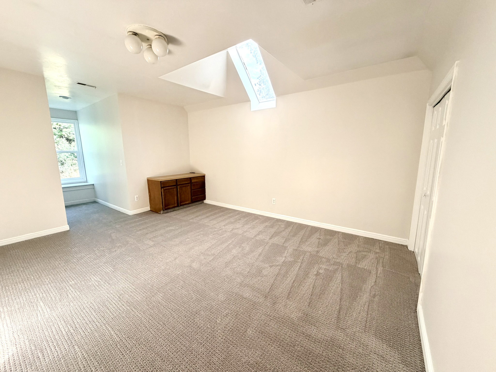 Spacious bedroom featuring neutral carpeting, a large window with natural light, a built-in wooden cabinet, and a ceiling-mounted light fixture