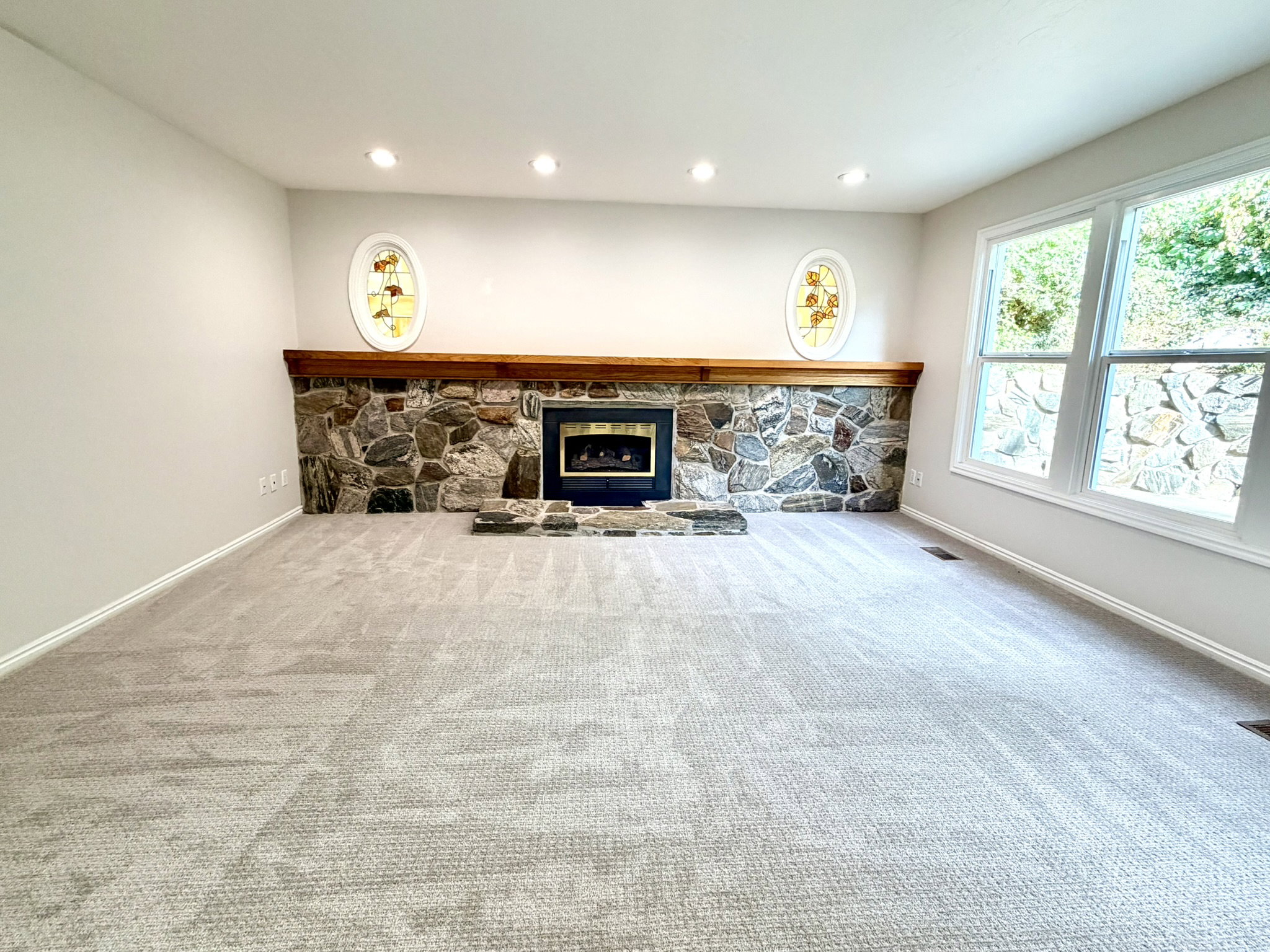 Spacious room featuring a stone-faced fireplace with a wood mantle, light-toned carpeting, recessed ceiling lighting, and large windows