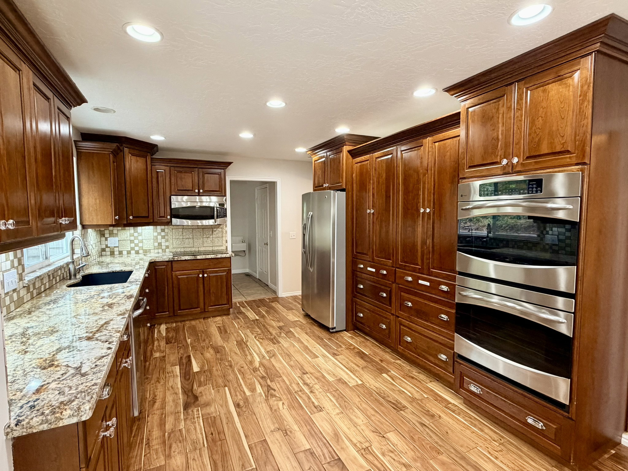 Gourmet kitchen featuring extensive wood cabinetry, granite countertops, stainless steel appliances including double ovens and a refrigerator, a tile backsplash, and wood-finish flooring