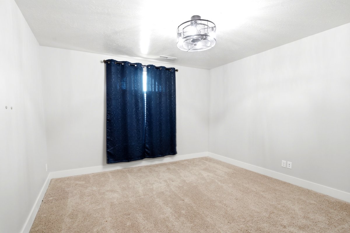 Neutral-toned room featuring a window with dark curtains, light-colored carpeting, white baseboards, and a contemporary ceiling fan
