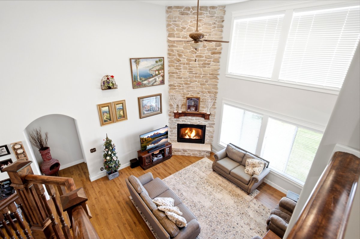 Vaulted ceiling living area featuring a floor-to-ceiling stone-faced fireplace with wood mantel, a ceiling fan, wood-finish flooring, and multiple large windows
