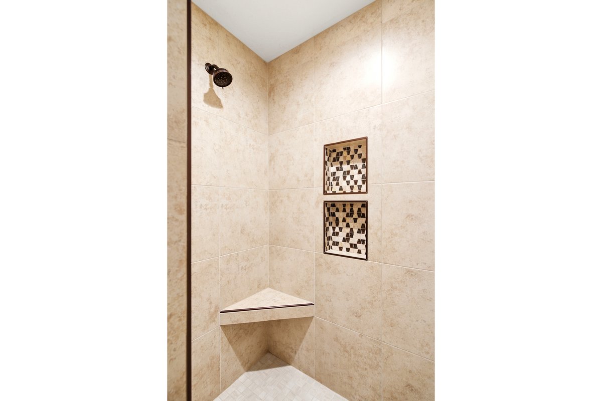 Tiled shower enclosure featuring a bronze showerhead, two recessed shelving niches with decorative mosaic tile accents, and a corner bench seat