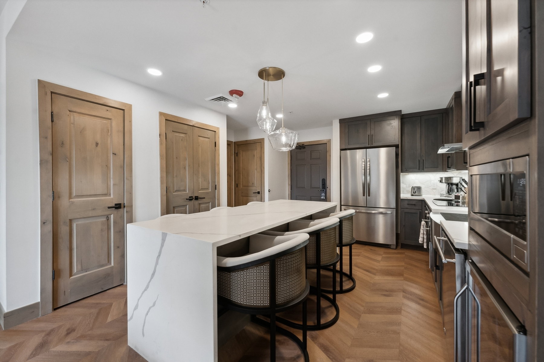 Kitchen featuring a waterfall-edge island with seating, herringbone wood-finish flooring, dark wood cabinetry, stainless steel appliances, and recessed lighting