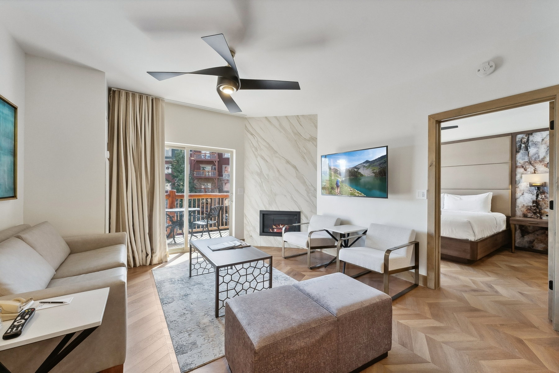 Living area featuring herringbone wood-finish flooring, a modern ceiling fan, a fireplace with a light-toned stone surround, and a sliding glass door opening to an outdoor patio with a wooden railing