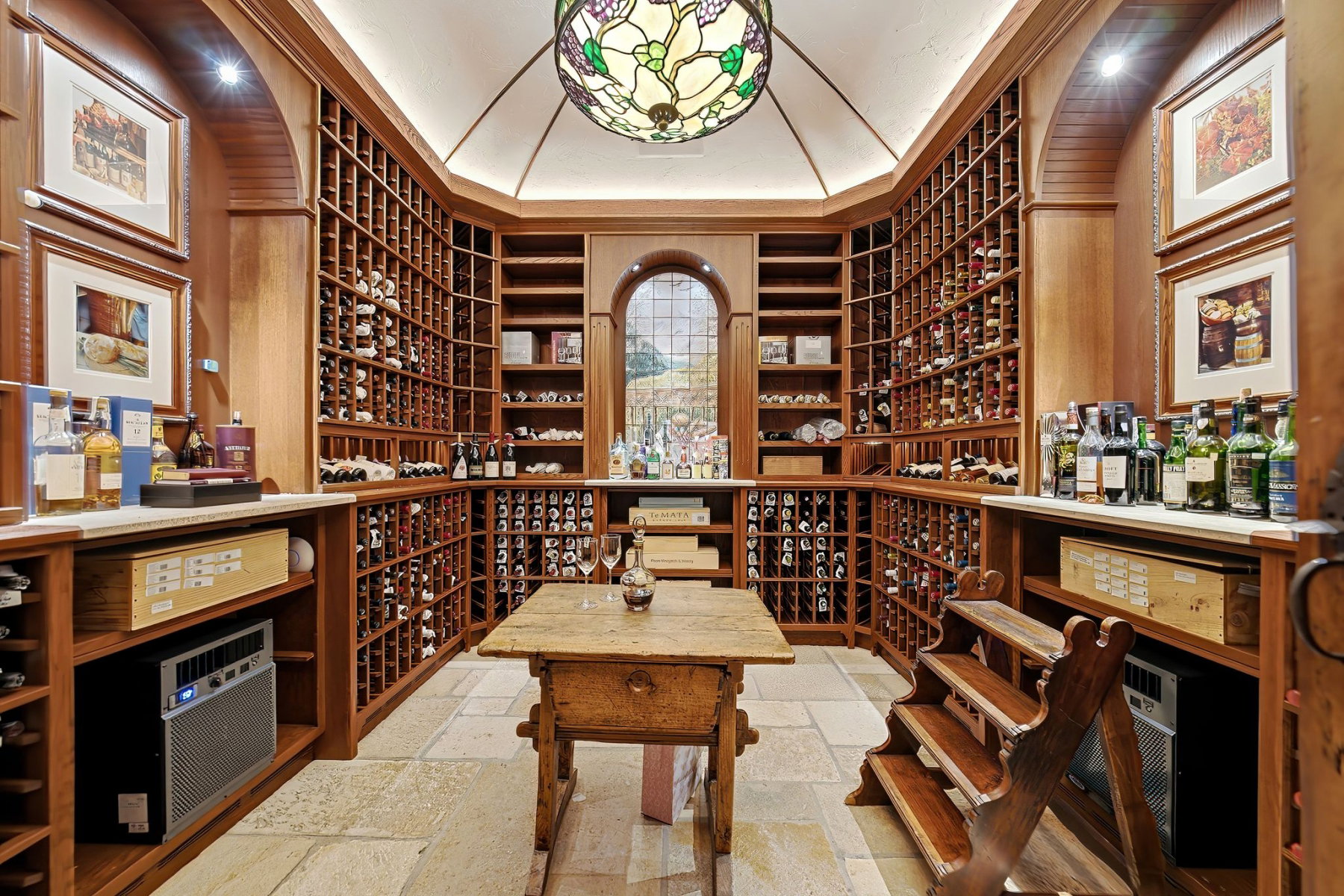 Dedicated wine cellar featuring extensive built-in wooden racking, stone tile flooring, a central wooden table, a stained-glass ceiling fixture, and a large arched window