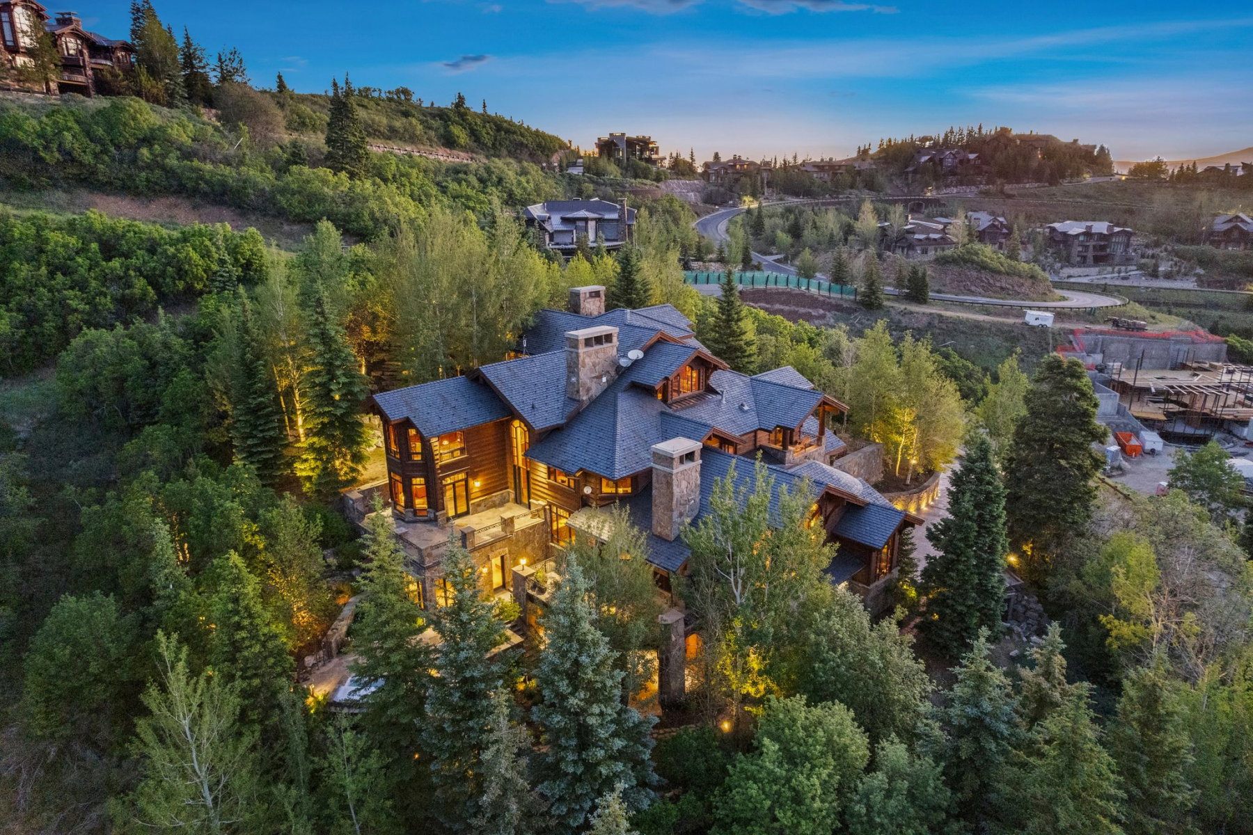 Hillside property featuring a multi-level structure with stone and wood siding, multiple chimneys, a dark shingle roof, and ample exterior lighting