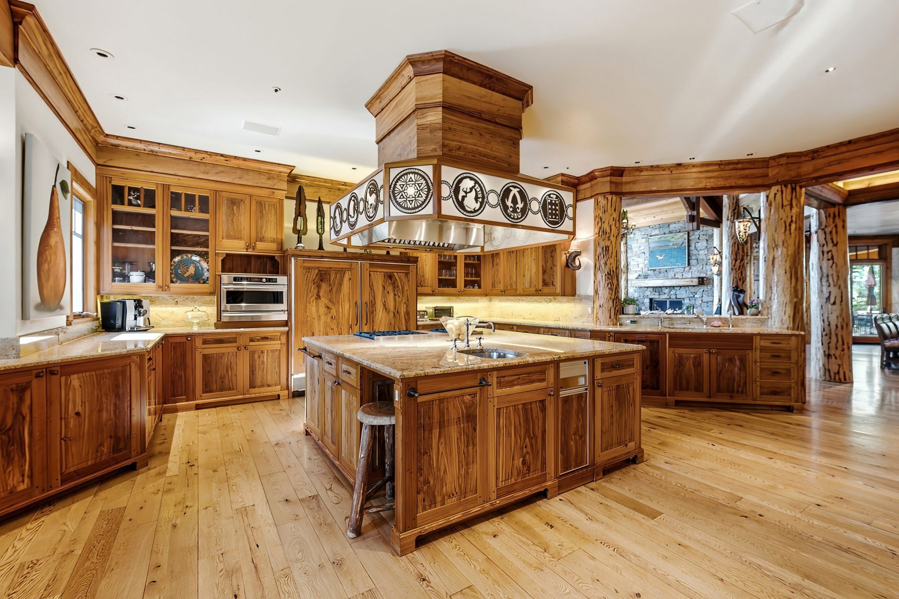 Gourmet kitchen with wood-finish flooring, extensive natural wood cabinetry, light-toned stone countertops, a large center island with integrated sink, and stainless steel appliances