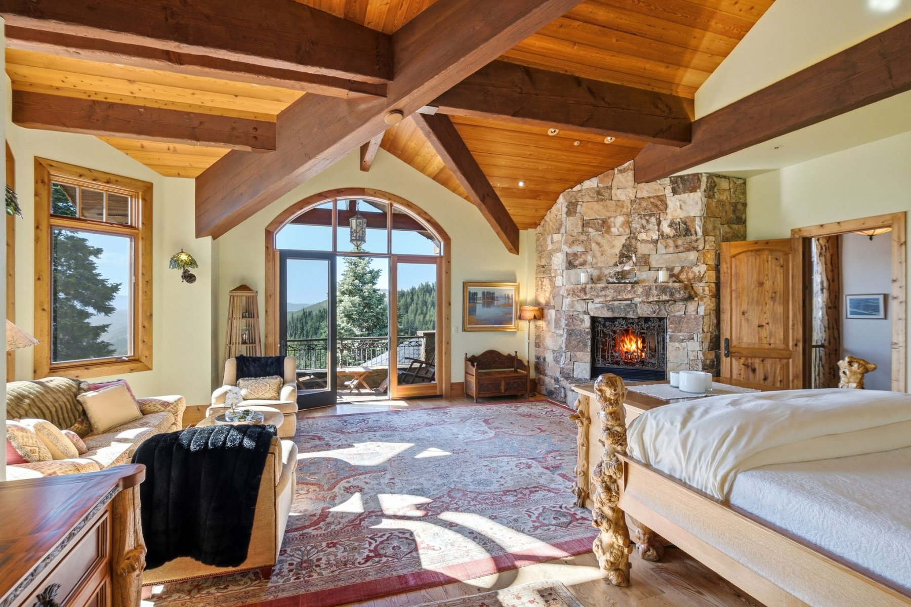 Spacious room featuring a stone-clad fireplace, exposed wood beams, vaulted wood-finish ceilings, an arched glass door, and large windows