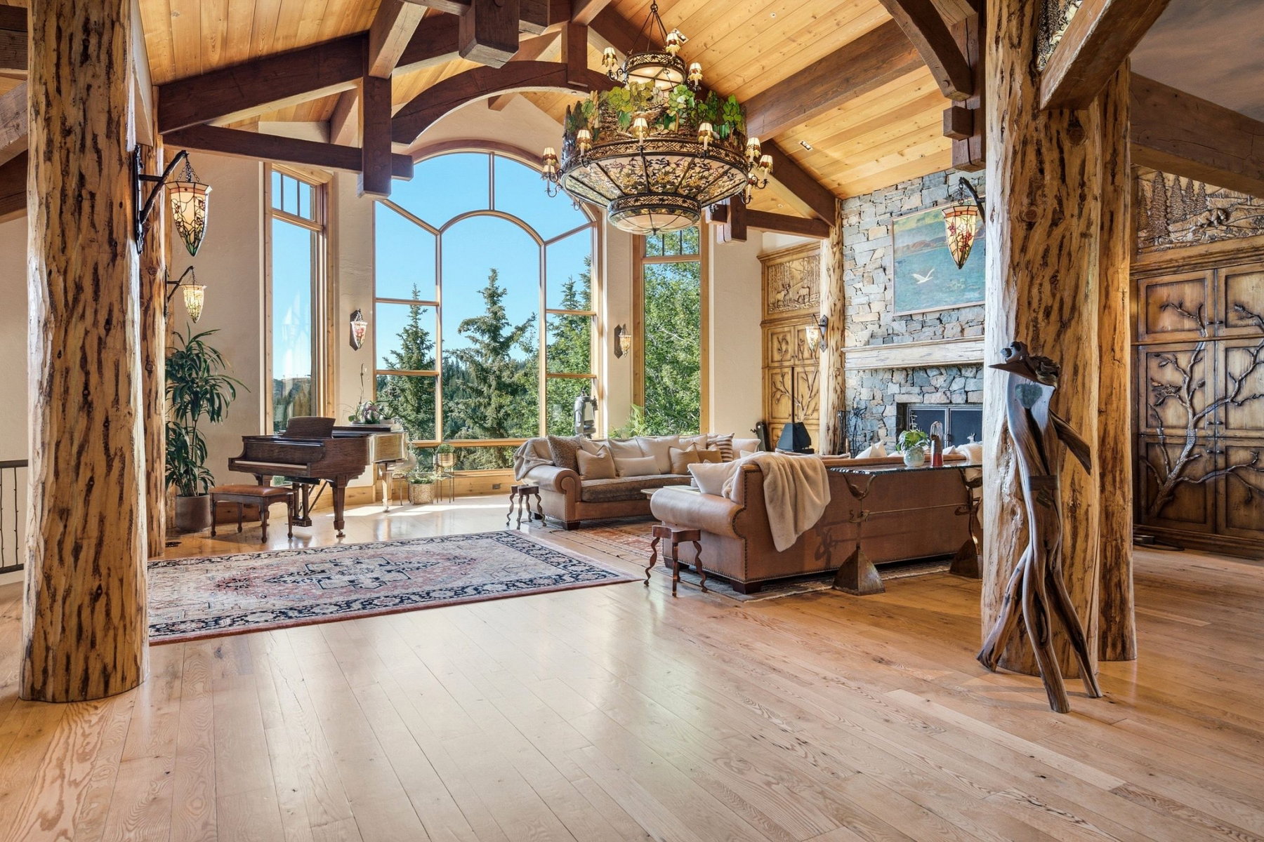 Grand living space featuring expansive arched windows, natural wood-finish flooring, exposed timber beams, a stone-clad fireplace, and rustic carved wooden doors