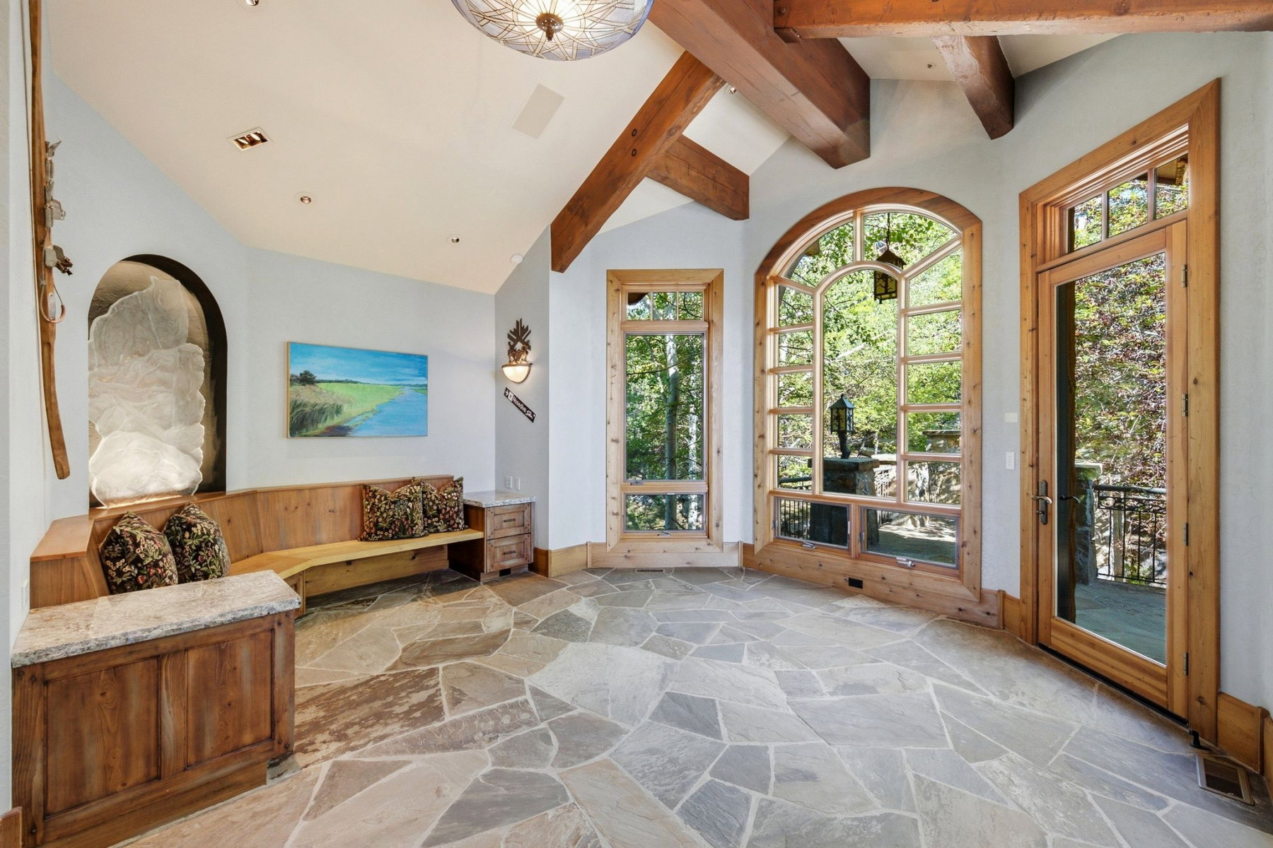 Spacious room featuring irregular flagstone flooring, exposed timber ceiling beams, an arched window with wooden framing, a wooden built-in bench, and a glass-paneled door